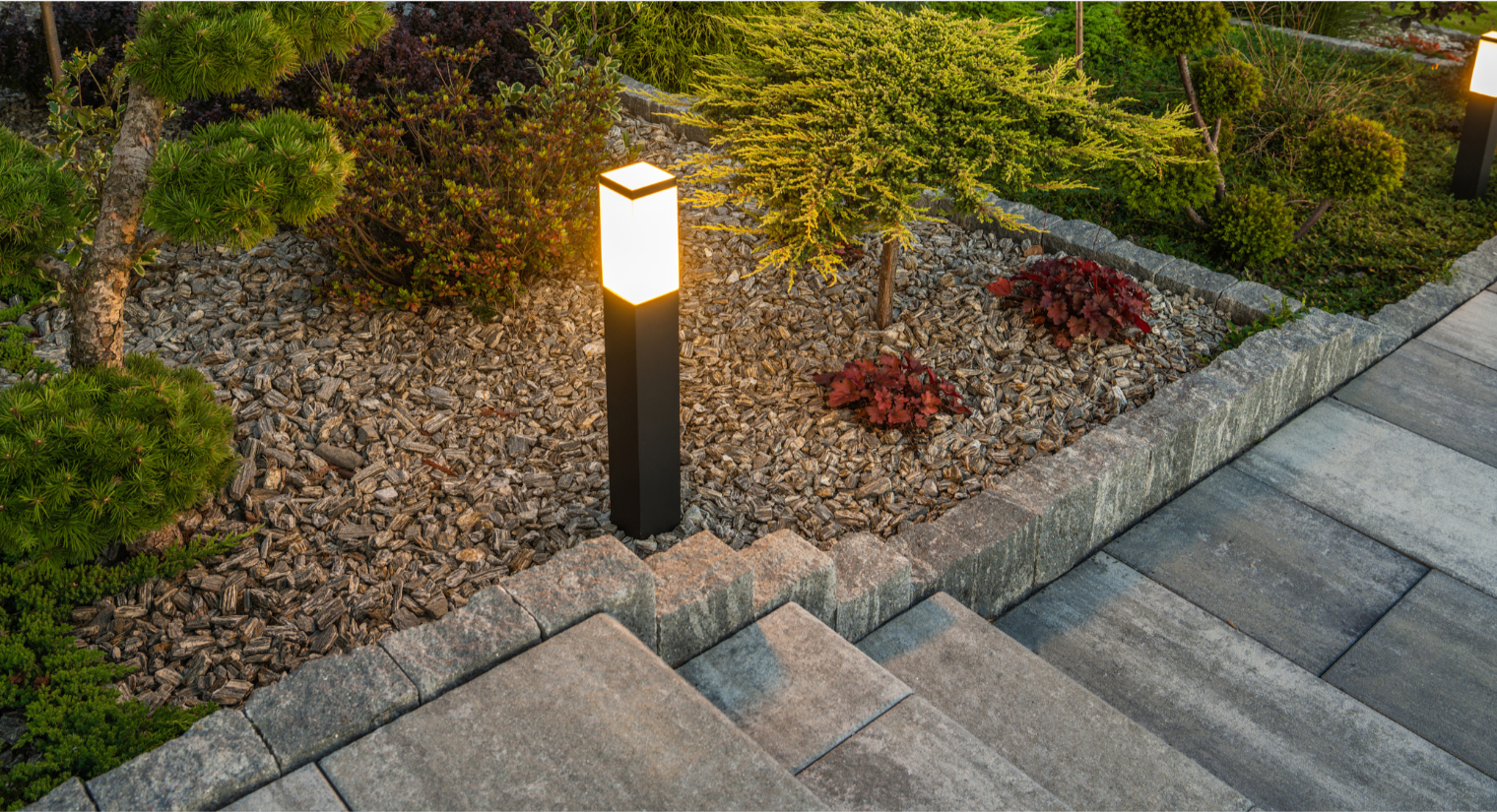 Nighttime landscape with a lit modern outdoor lamp post illuminating a garden bed with small bushes, trees, and red and green foliage, bordered by stone pavers and a stone edging.