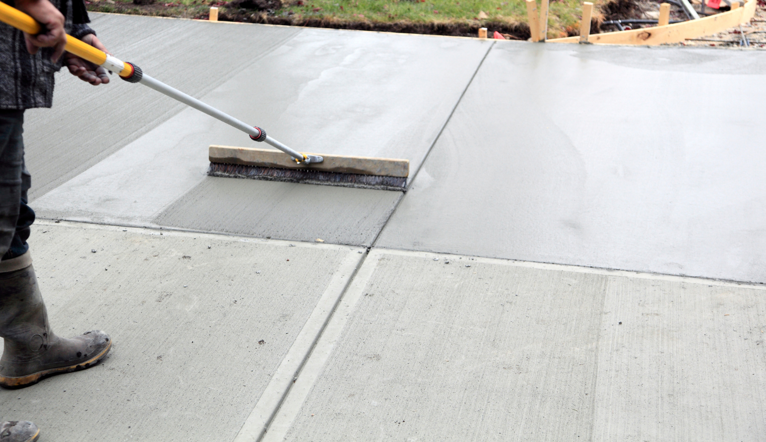 Person using a long-handled tool to smooth wet concrete on a sidewalk or driveway during construction.