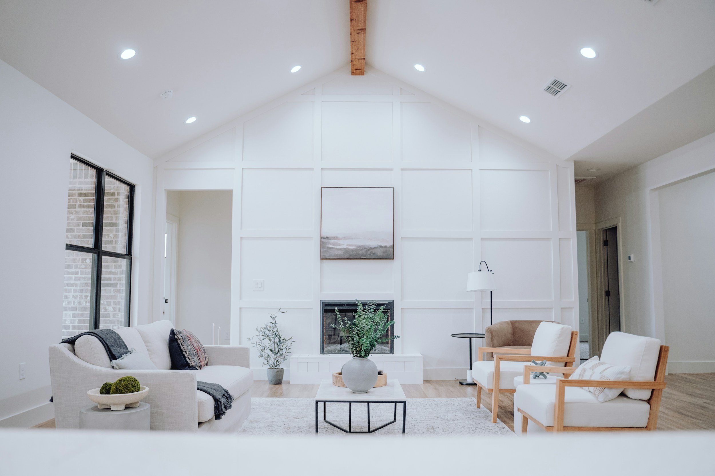 Bright living room with white walls, high vaulted ceiling with wooden beam, featuring a large window on the left, a fireplace, and modern furniture including a beige sofa, wooden armchairs, a black coffee table with plants, and a floor lamp.