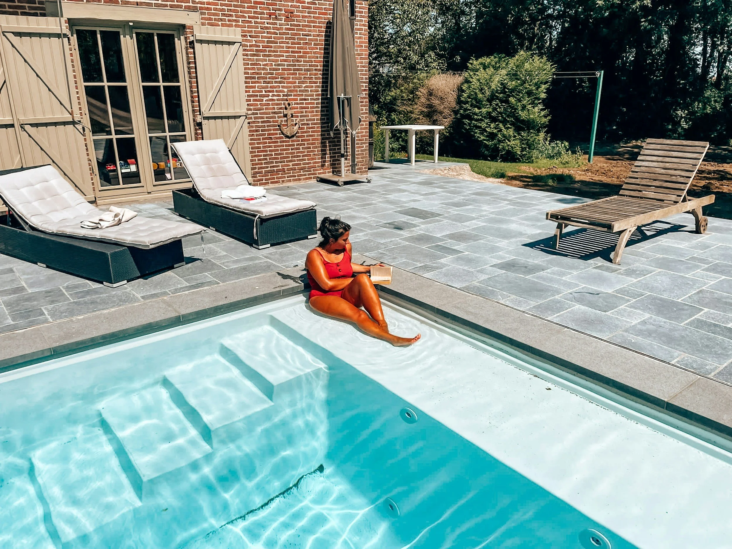 A woman in a red swimsuit relaxing by a swimming pool, reading a book, with poolside lounge chairs, a brick house wall, and greenery in the background.