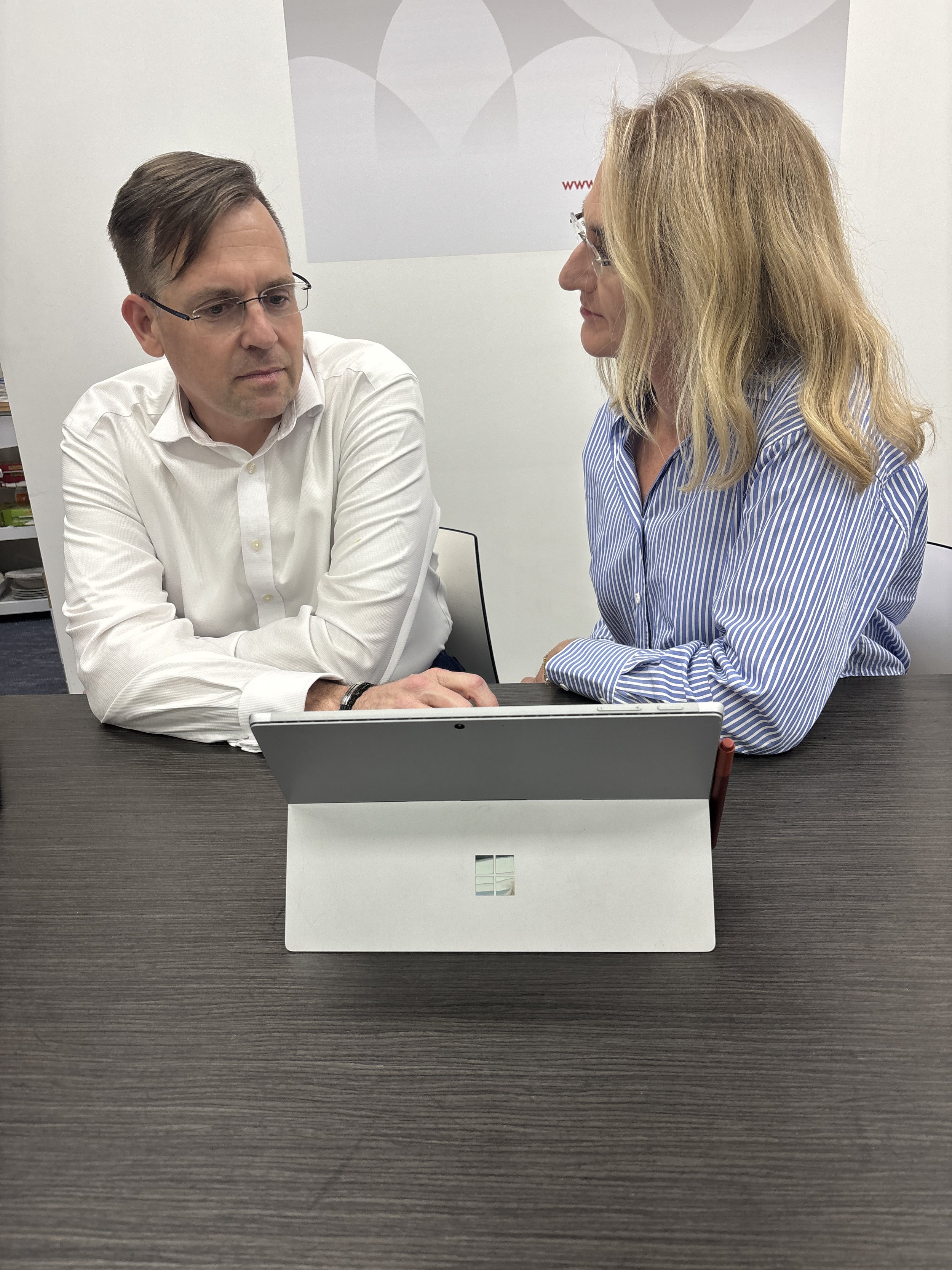 Two people sitting at a conference table, engaged in conversation, with a Microsoft Surface device on the table in front of them.