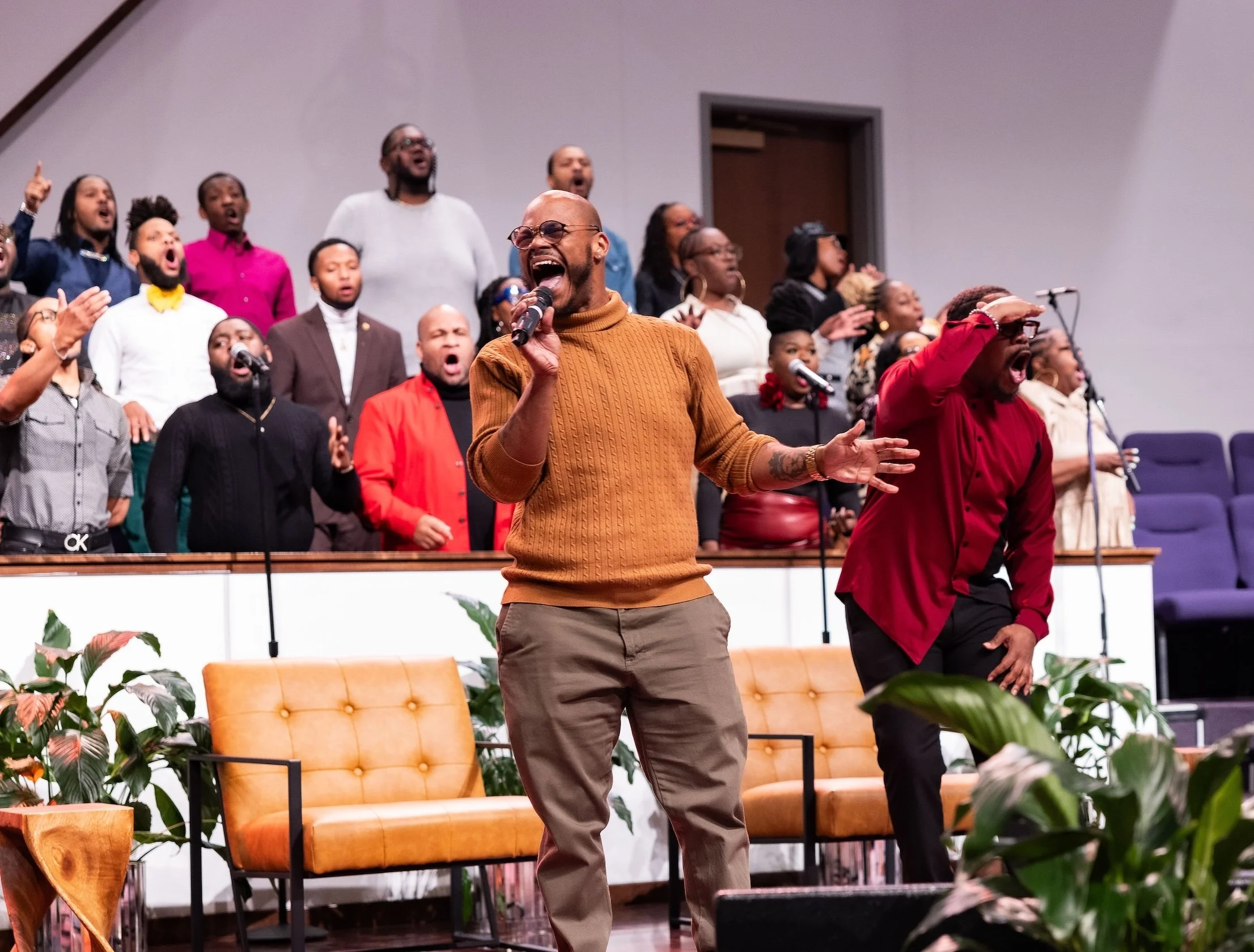 A group of people singing and performing on stage, with two men in front holding microphones, one in an orange sweater and the other in a red shirt, surrounded by plants and chairs.