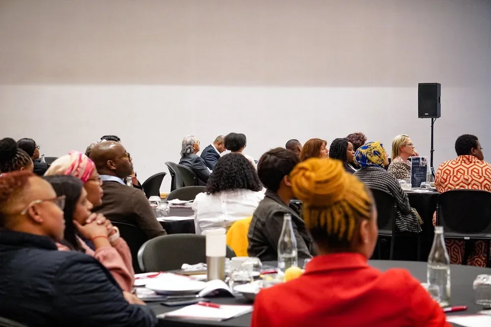 Audience seated at round tables listening to a presentation in a conference room.