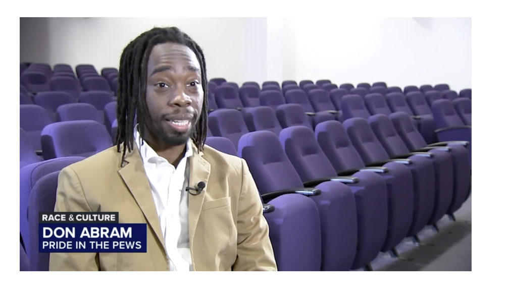 Man with dreadlocks sitting in a mostly empty auditorium with purple seats, speaking during an interview.