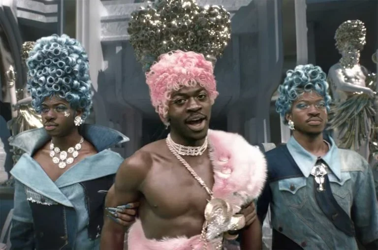 Three men in extravagant, colorful costumes with wigs, jewelry, and dramatic makeup, standing in front of a decorative background with mannequins and Christmas decorations.