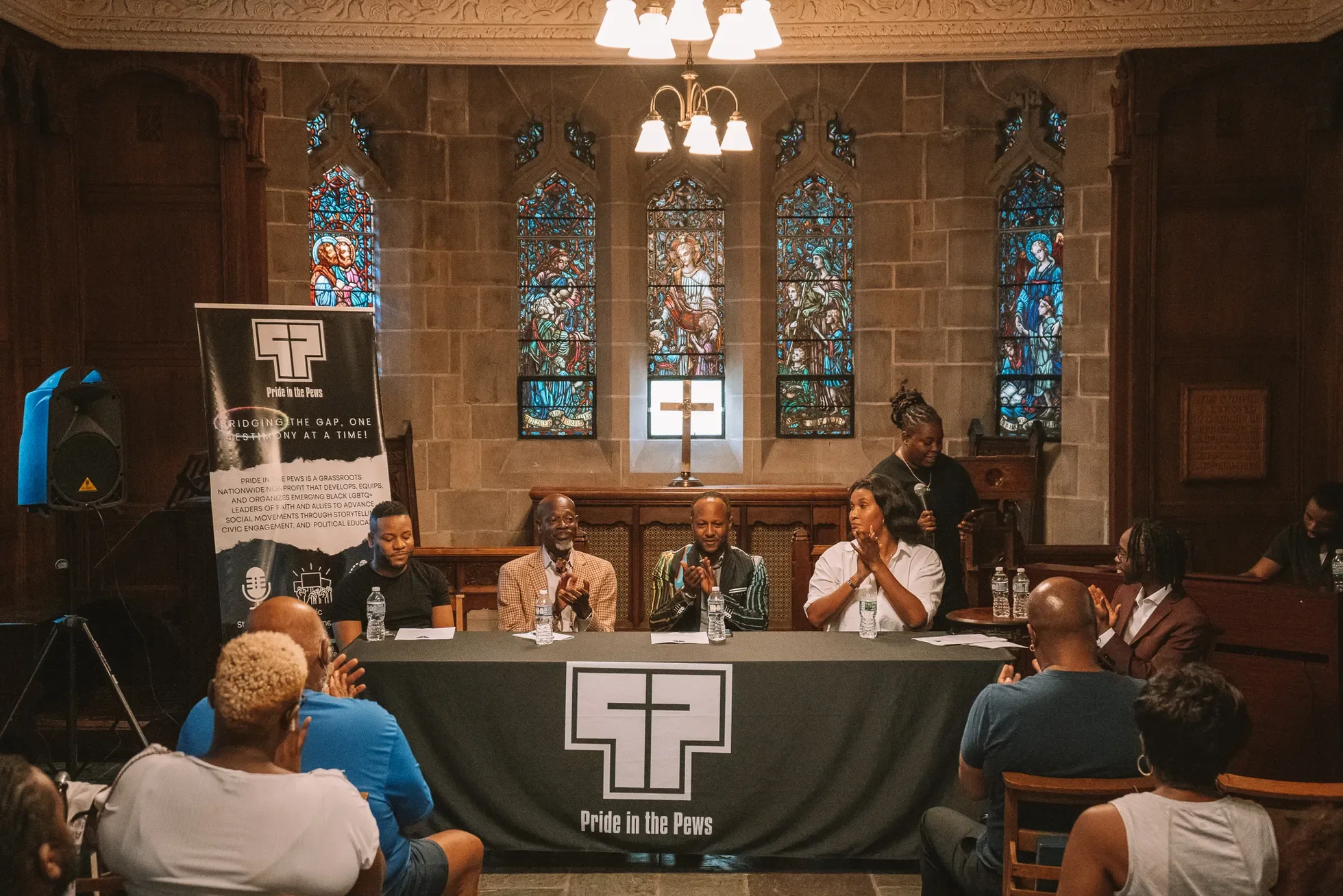 A panel of six people sitting at a long table in a church with stained glass windows behind them. The table has a banner with a cross and the text "Pride in the Pews." One person is speaking into a microphone, while others are clapping or listening, with audience members seated in front.