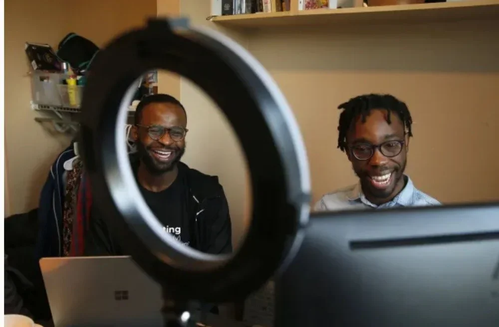 Two men with glasses smiling and recording a video or making a video call, viewed through a camera lens.