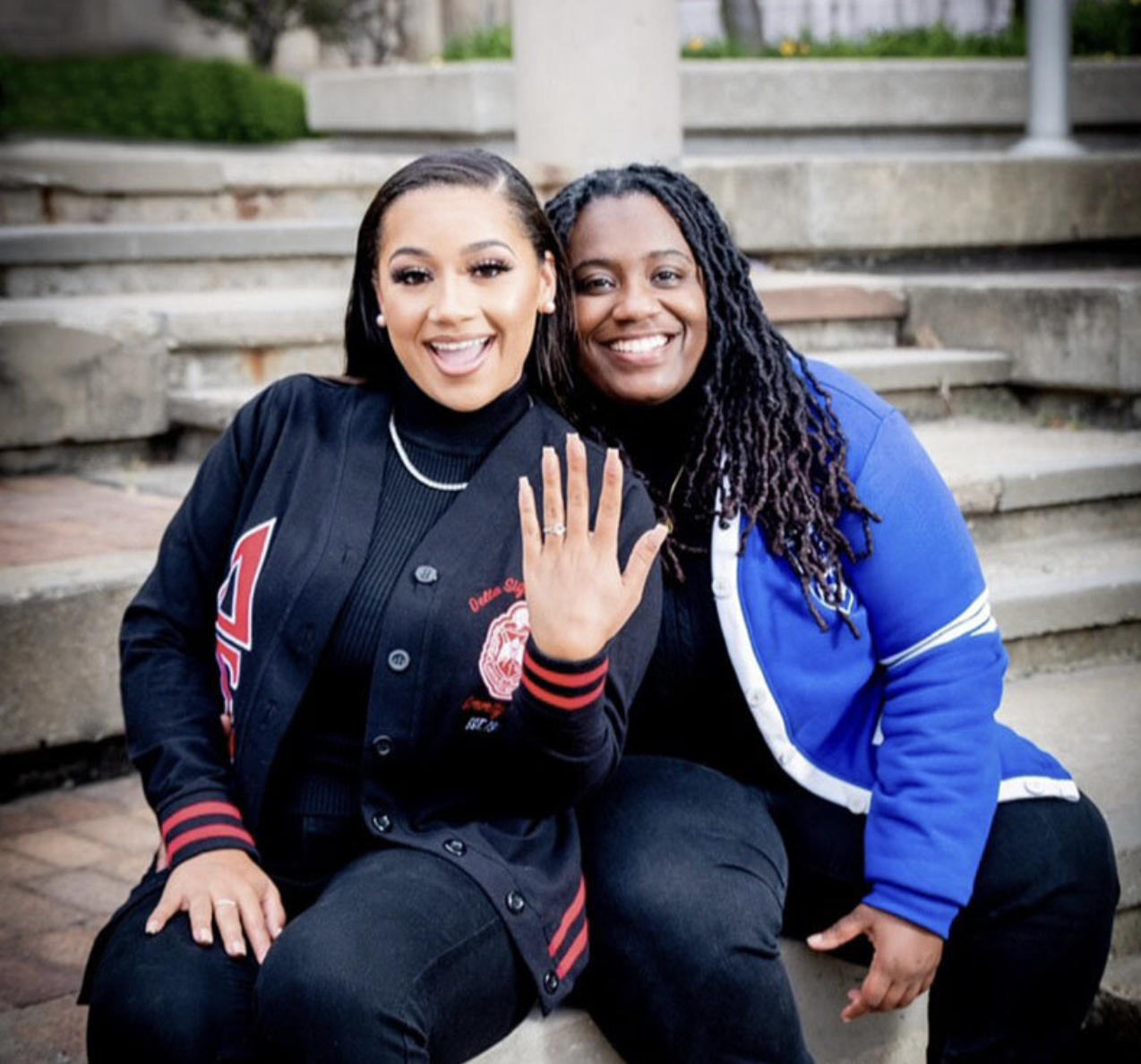 Two women sitting on outdoor stairs, smiling at the camera. The woman on the left is showing her engagement ring.
