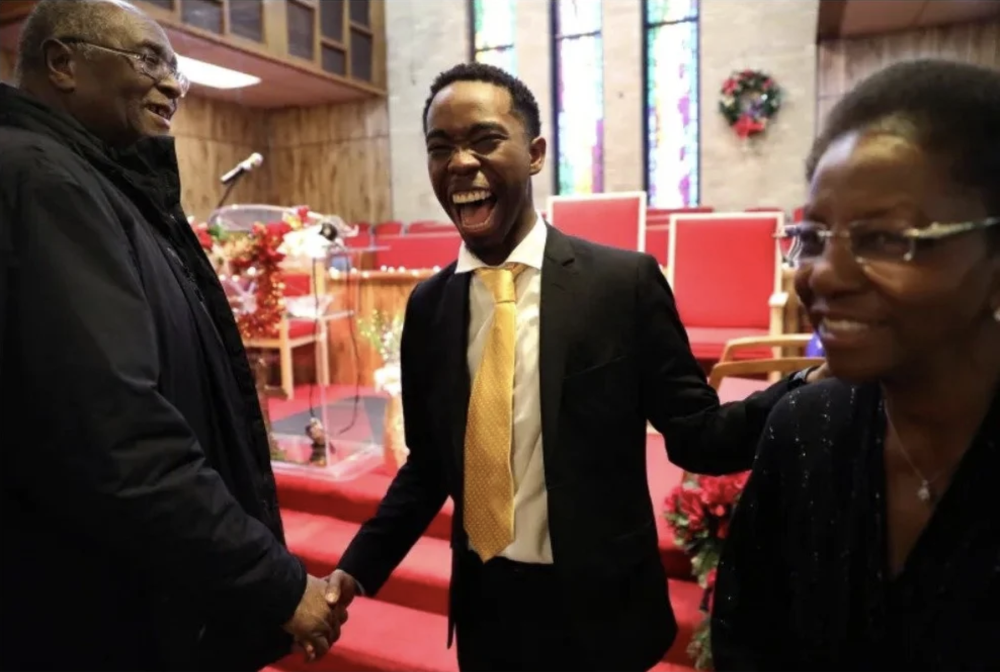 Three people smiling and shaking hands inside a church with red chairs, flowers, and stained glass windows in the background.