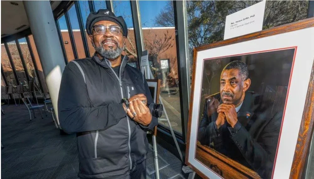 A man with glasses and a beard smiling, standing next to a portrait of another man with a serious expression, wearing a suit and resting his chin on his hands.