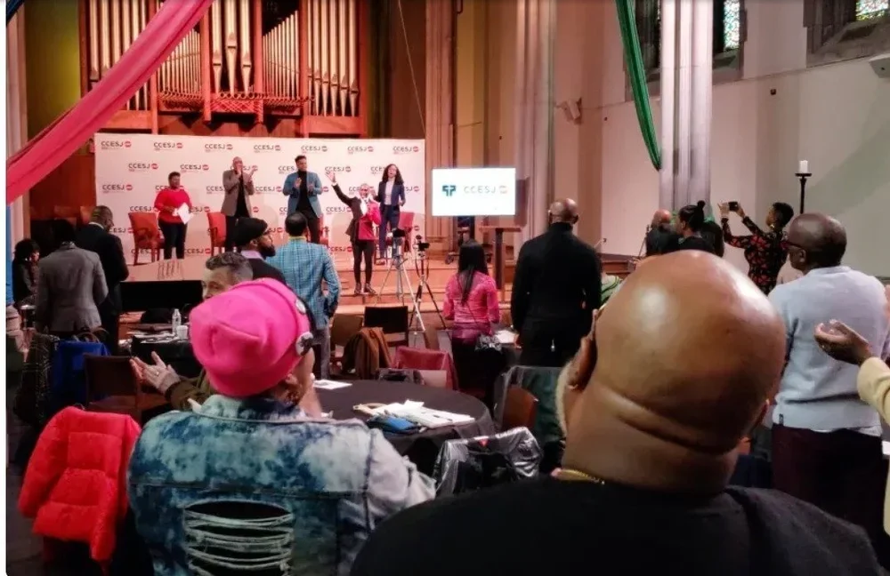 People attending a conference or event with a stage at the front, featuring four speakers and a large screen, in a room with wooden organ pipes and decorated with pink and green fabric.