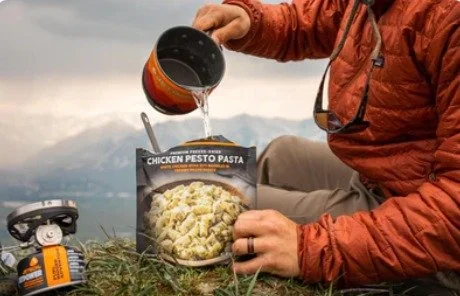 A person pouring water into a package of Peak ReFuel chicken pesto pasta while sitting outdoors with a mountain backdrop.