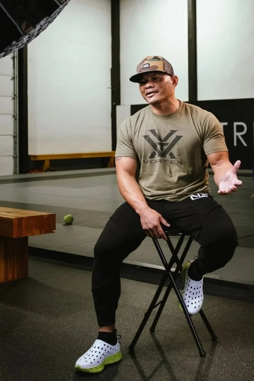 Wilton sitting on a chair in a gym, wearing a camouflage cap, a green T-shirt with a Vulkan logo, black pants, and white and yellow athletic shoes, gesturing with his right hand.