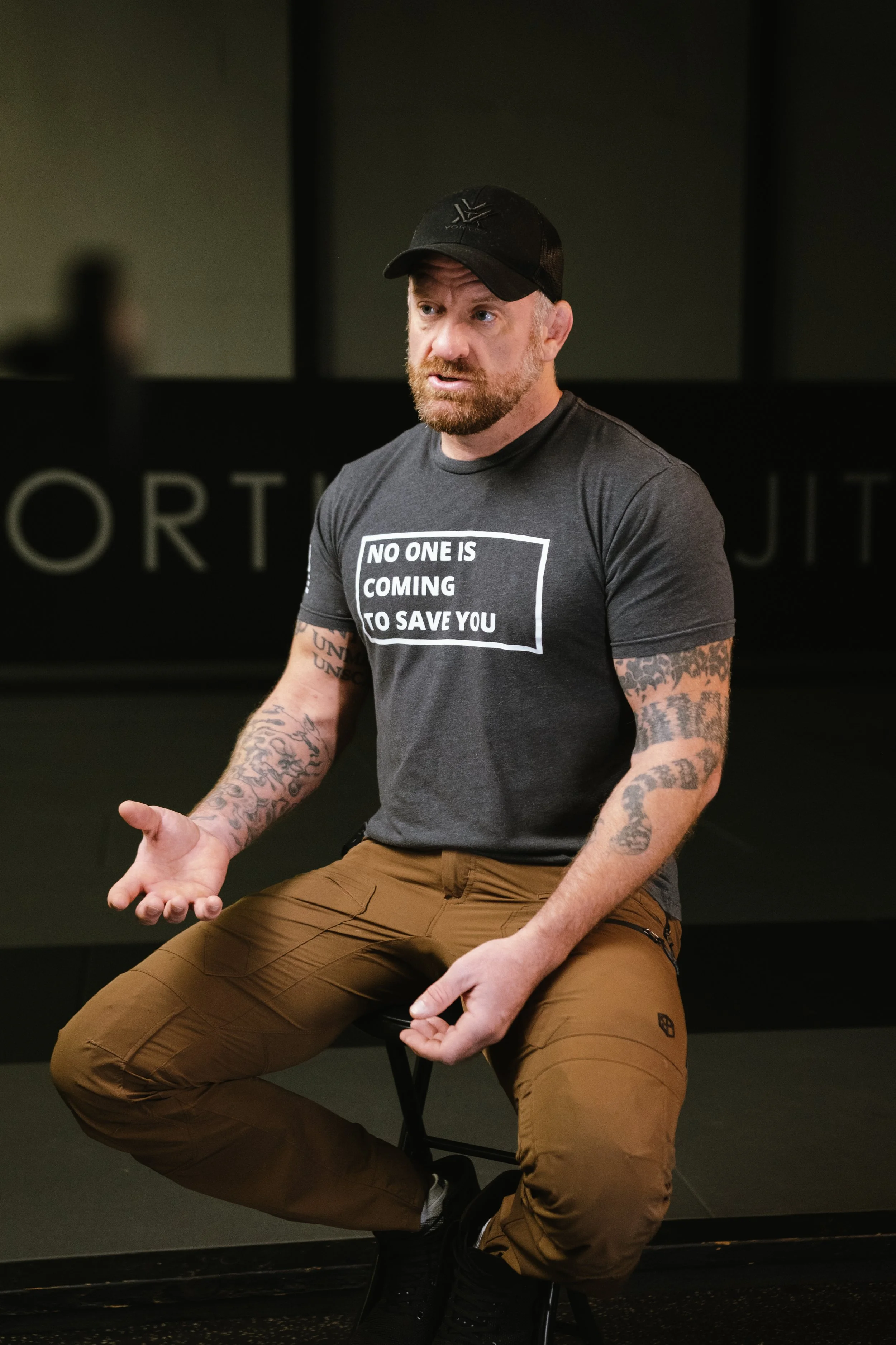 Greg with tattoos, wearing a black cap and gray t-shirt with the text 'NO ONE IS COMING TO SAVE YOU,' is sitting on a stool and speaking with expressive hand gestures in a gym or fitness studio.
