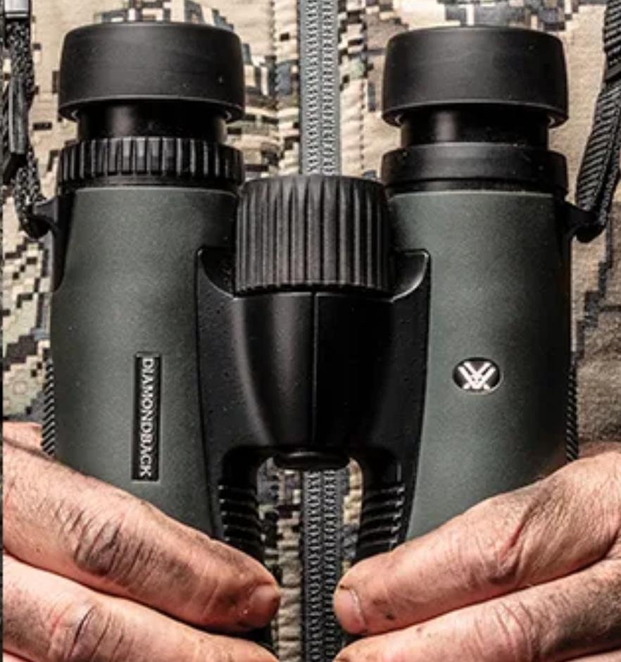 Close-up of a black pair of binoculars held by a person's hands, resting on a patterned surface from Vortex optics.