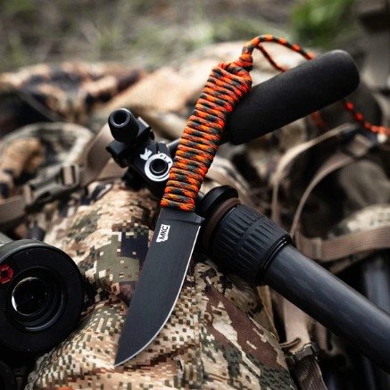A survival knife with a black blade and a black handle, attached to a strap with orange and black paracord, resting on camouflage fabric outdoors.