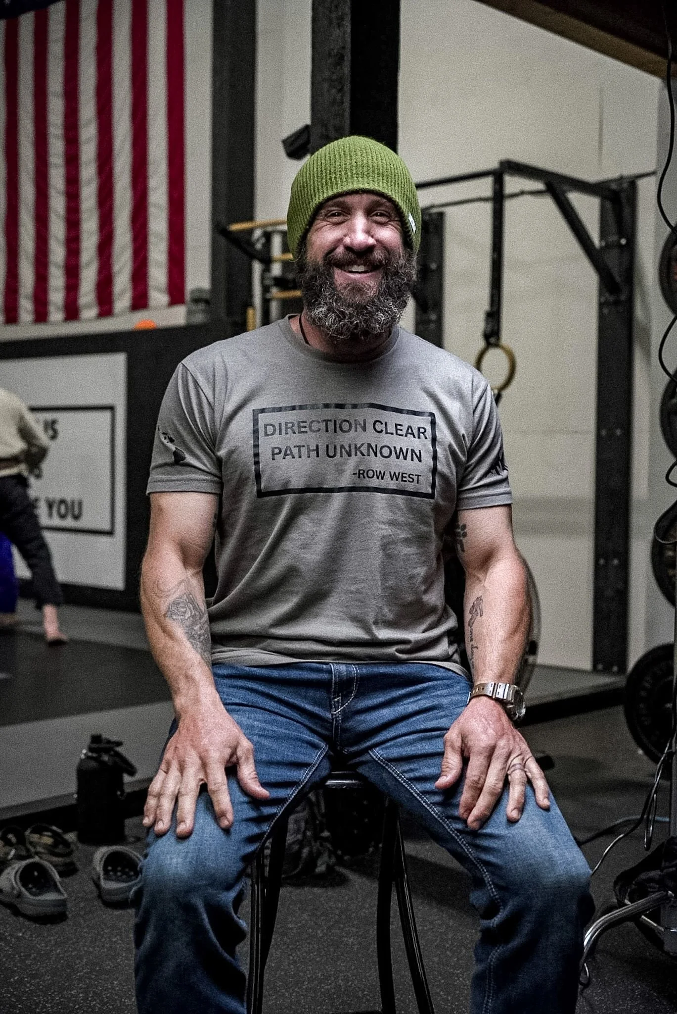 Joshua with a beard and a green knit beanie sitting on a stool in a gym, smiling at the camera. He is wearing a gray T-shirt with text and blue jeans, and has tattoos on his arms.