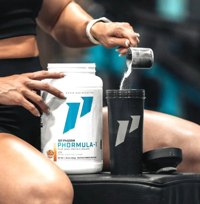 Person scooping powder from a container labeled 'Phormula-1' protein powder into a shaker bottle.