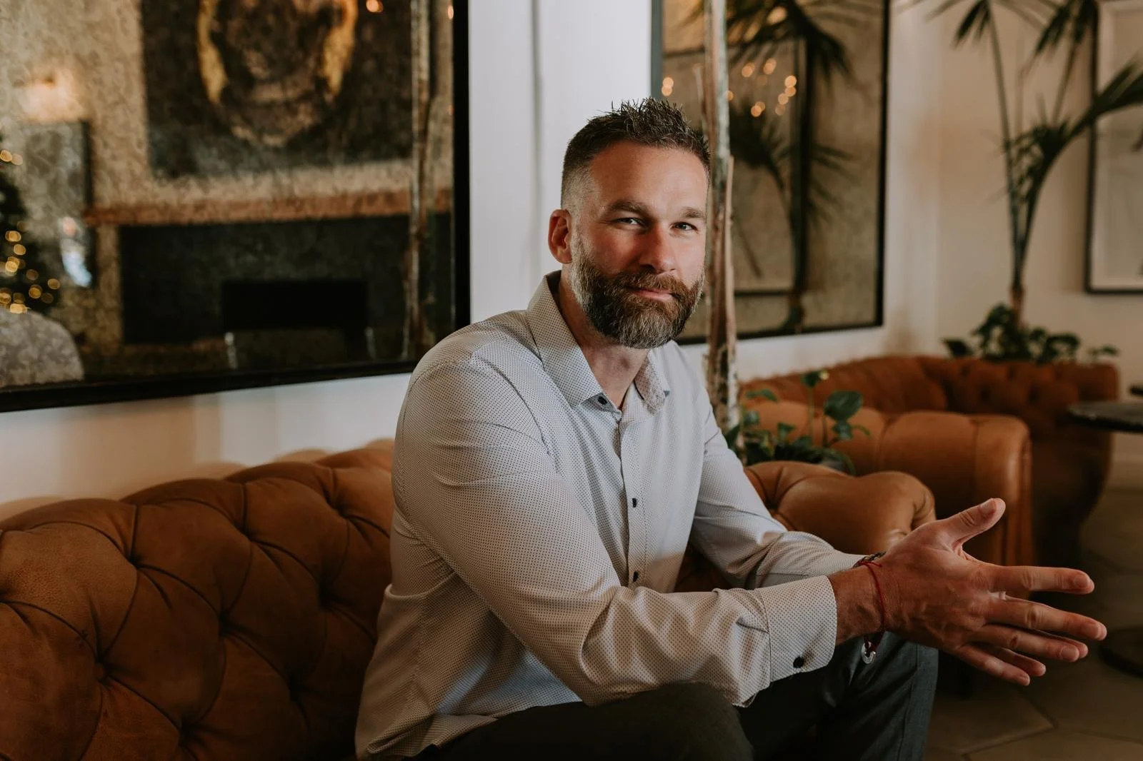 Jerremy Alexander Newsome with a beard and short hair, wearing a light gray button-up shirt, sitting on a brown leather couch in a cozy decorated room, smiling and gesturing with his hands.
