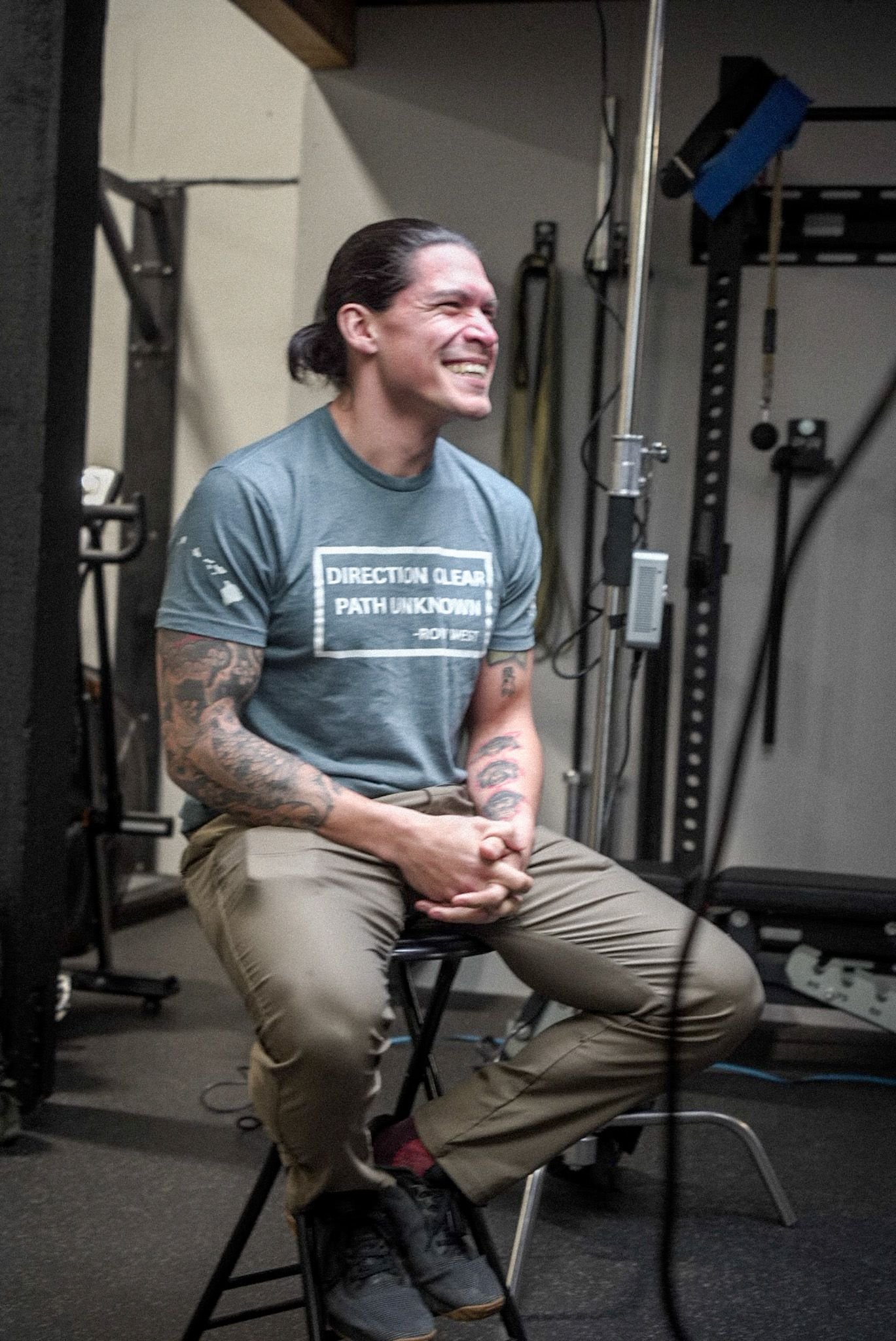 Johnny  with long dark hair and tattoos on his arms is sitting on a stool in a gym, smiling or laughing.