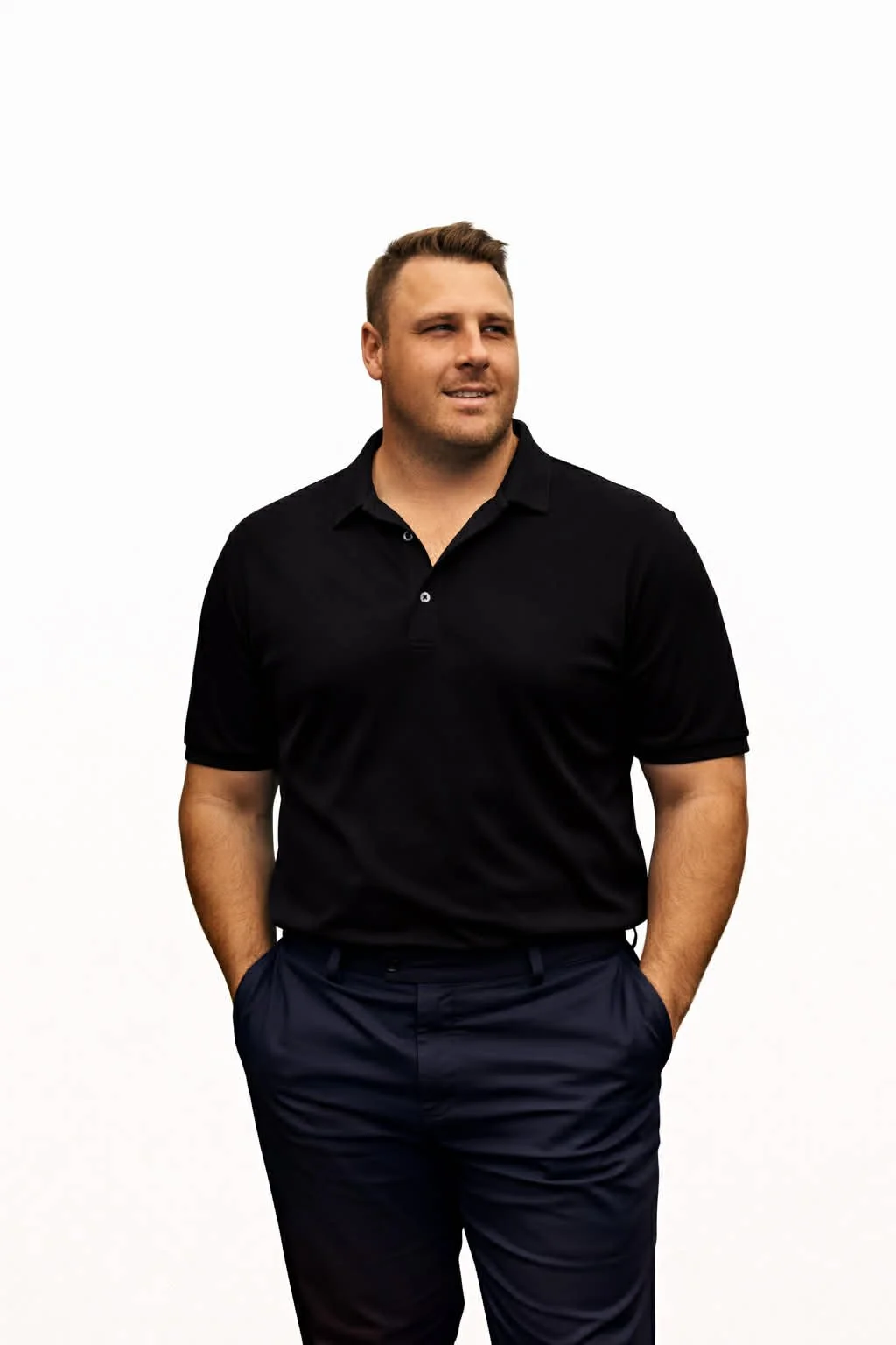 A young man with short hair, wearing a black polo shirt and navy pants, standing with his hands in his pockets against a white background.