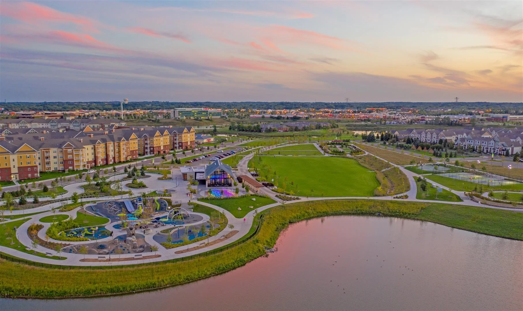 An aerial view of a park in Maple Grove, MN at sunset showing a pond, colorful playground, open grassy areas, walking paths, and apartment buildings in the background.