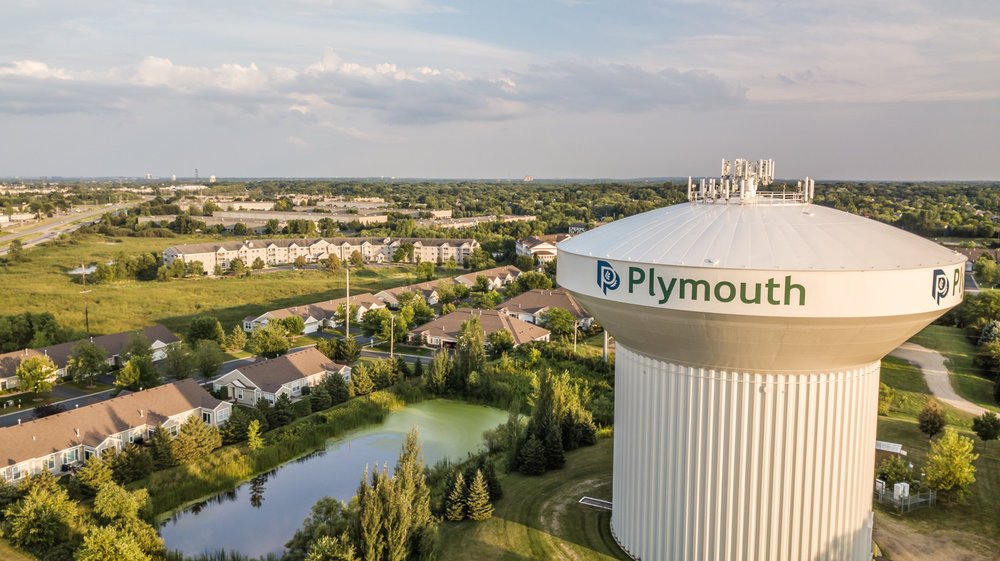 A large white water tower labeled 'Plymouth' surrounded by residential neighborhoods, trees, and a pond, with a partly cloudy sky in the background.
