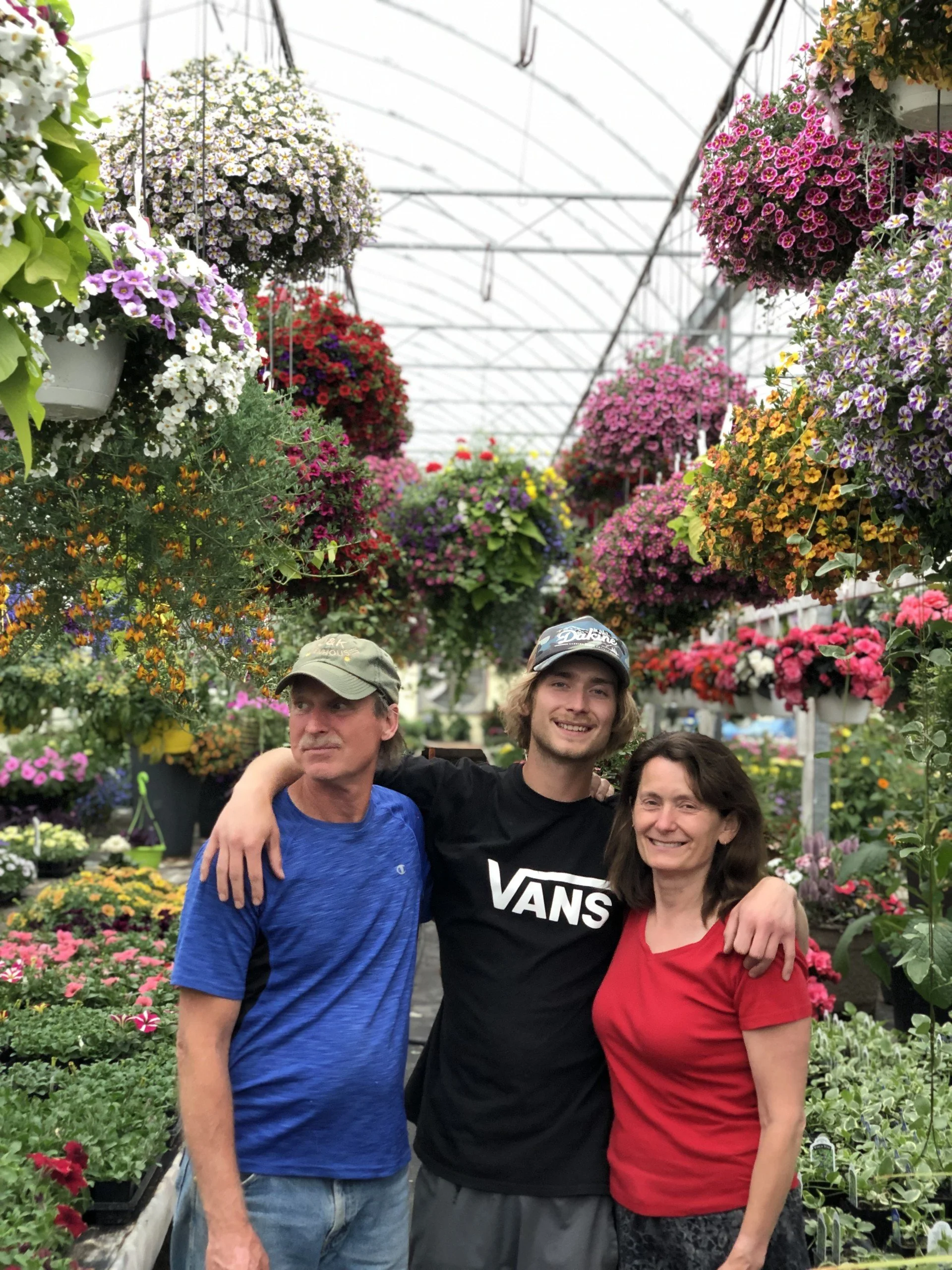Three people, two men and one woman, stand together inside a greenhouse filled with colorful hanging and potted flowers. They are smiling and have their arms around each other.