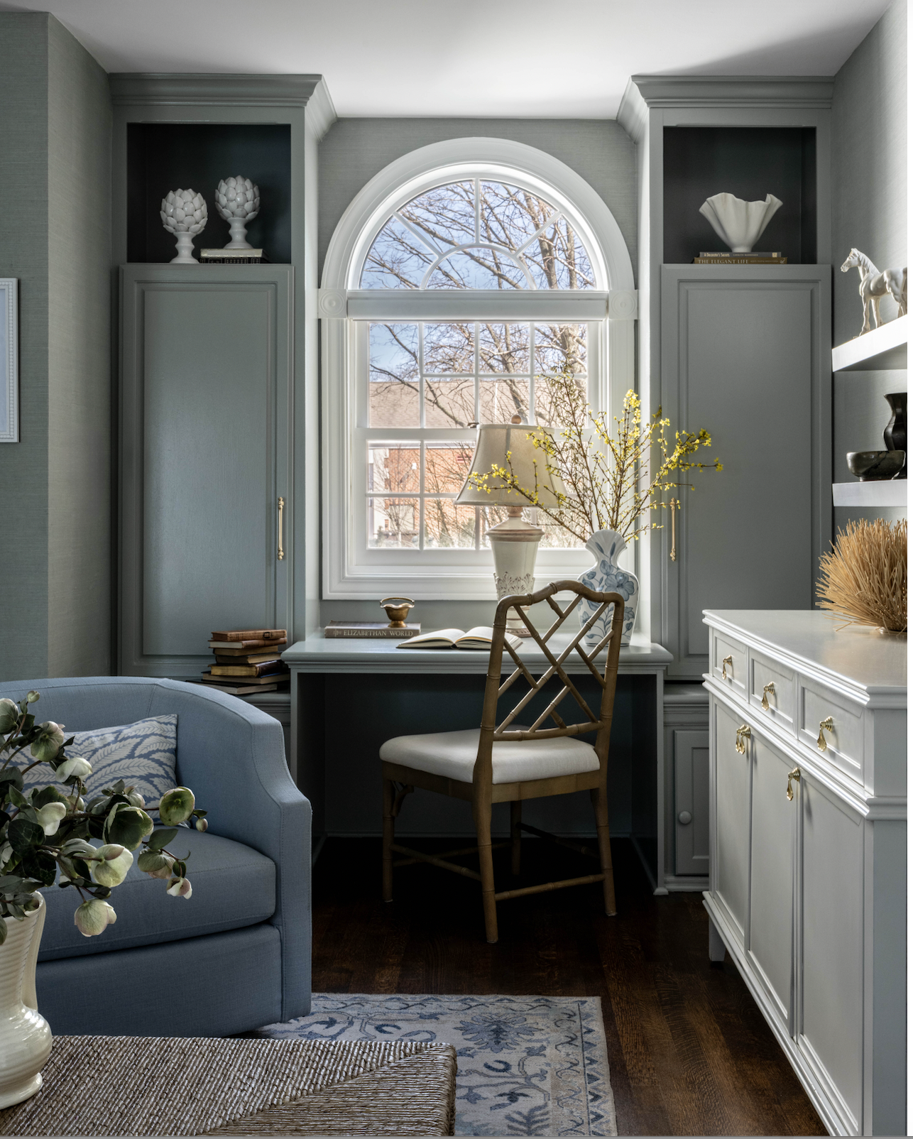Interior of a home office with a large arched window, a desk with a lamp and decorative vases, a wooden chair, a blue armchair, and a white cabinet with gold handles, decorated with plants and books.