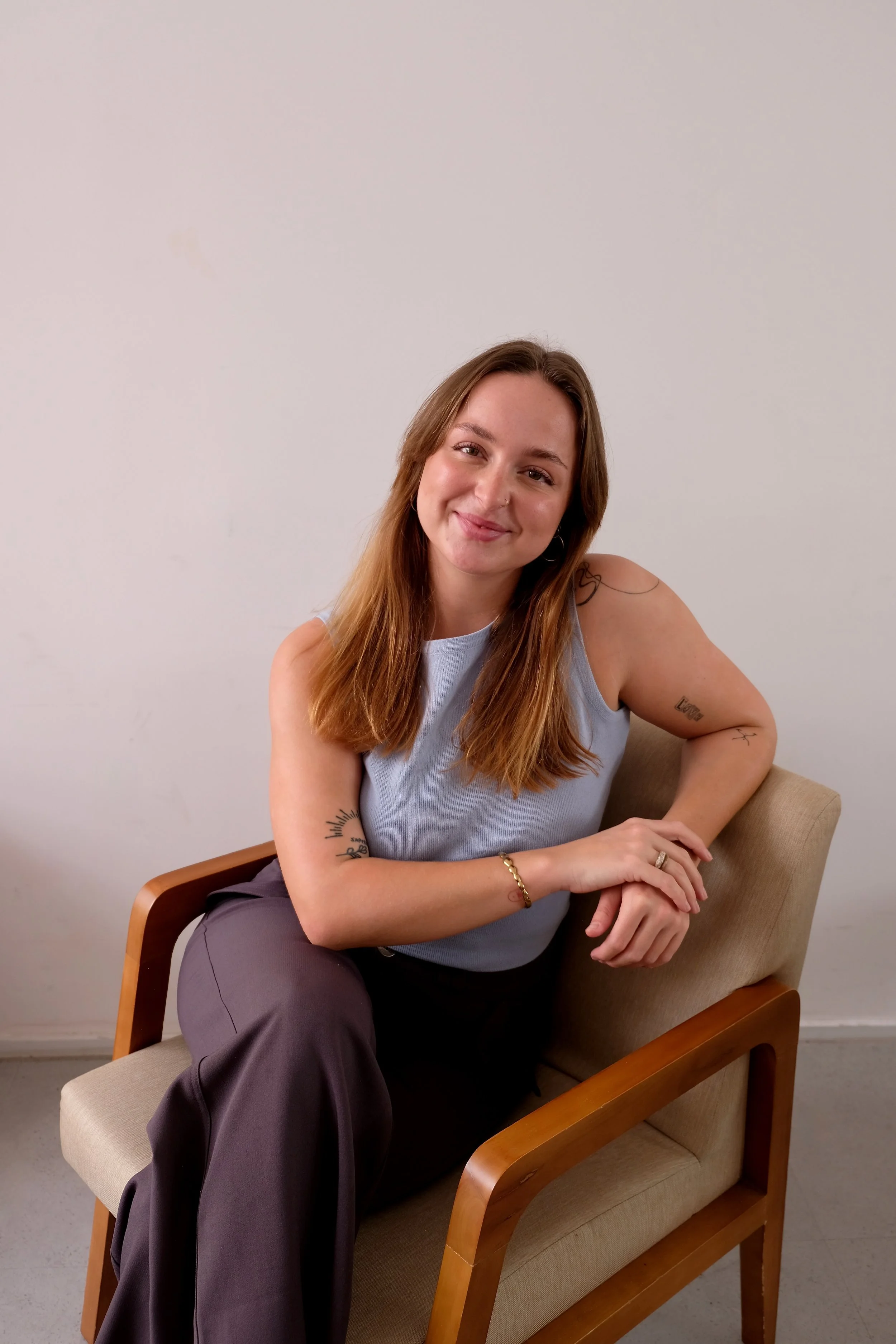 A woman with brown hair, wearing a light blue sleeveless top and dark pants, sitting on a beige armchair with wooden armrests, smiling at the camera against a plain white wall.