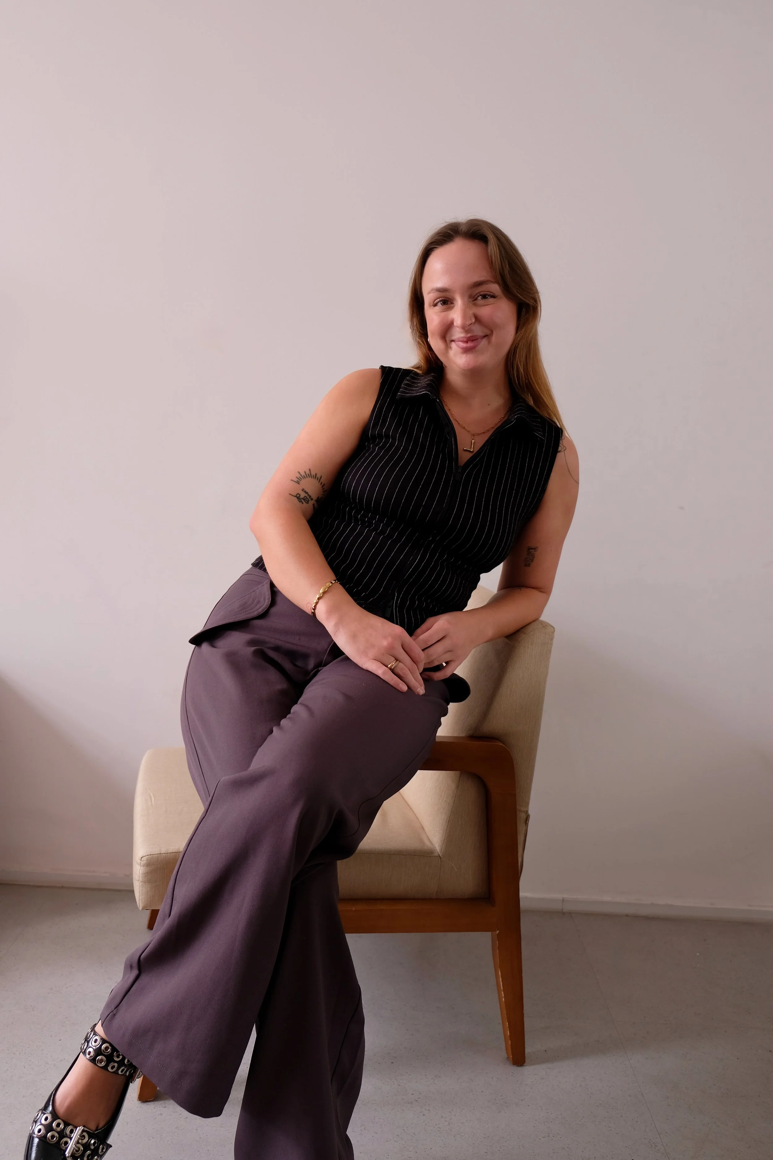 A woman sitting on a wooden chair with a beige cushion, wearing a black striped sleeveless top and dark wide-leg pants, smiling at the camera.
