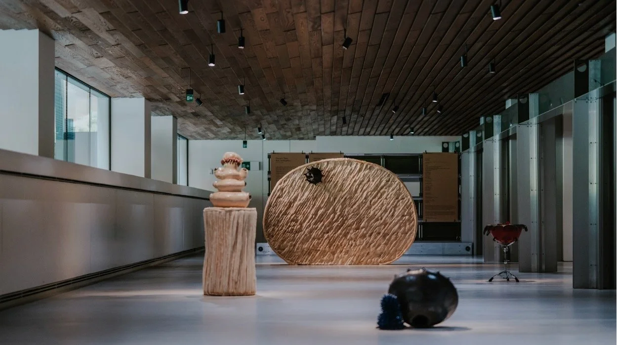 Modern art installation featuring wooden sculptures and objects in a spacious, well-lit room with a wooden ceiling and large windows.