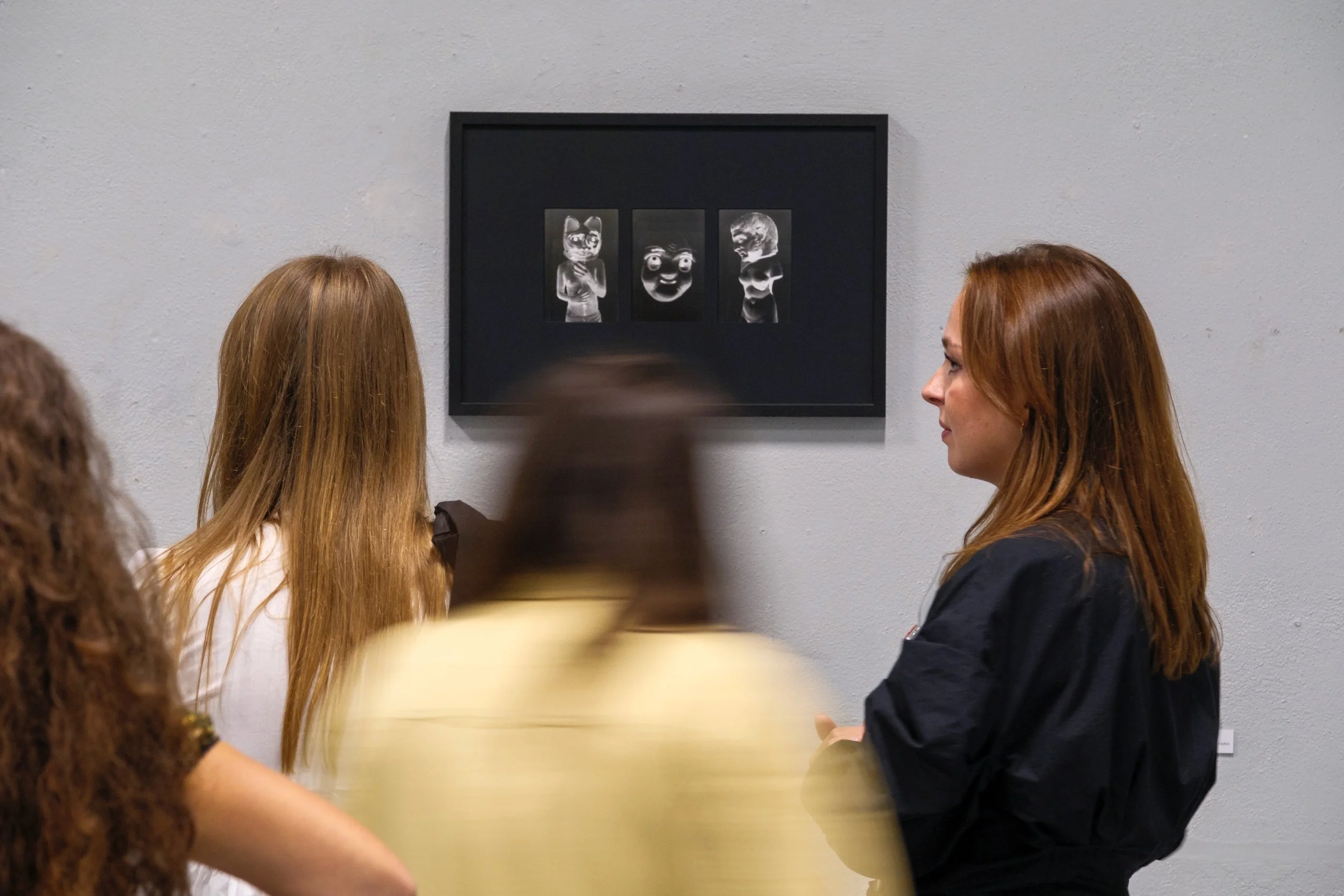 Art gallery with a black-framed artwork displaying four black and white character images on a gray wall, viewed by three women observing.