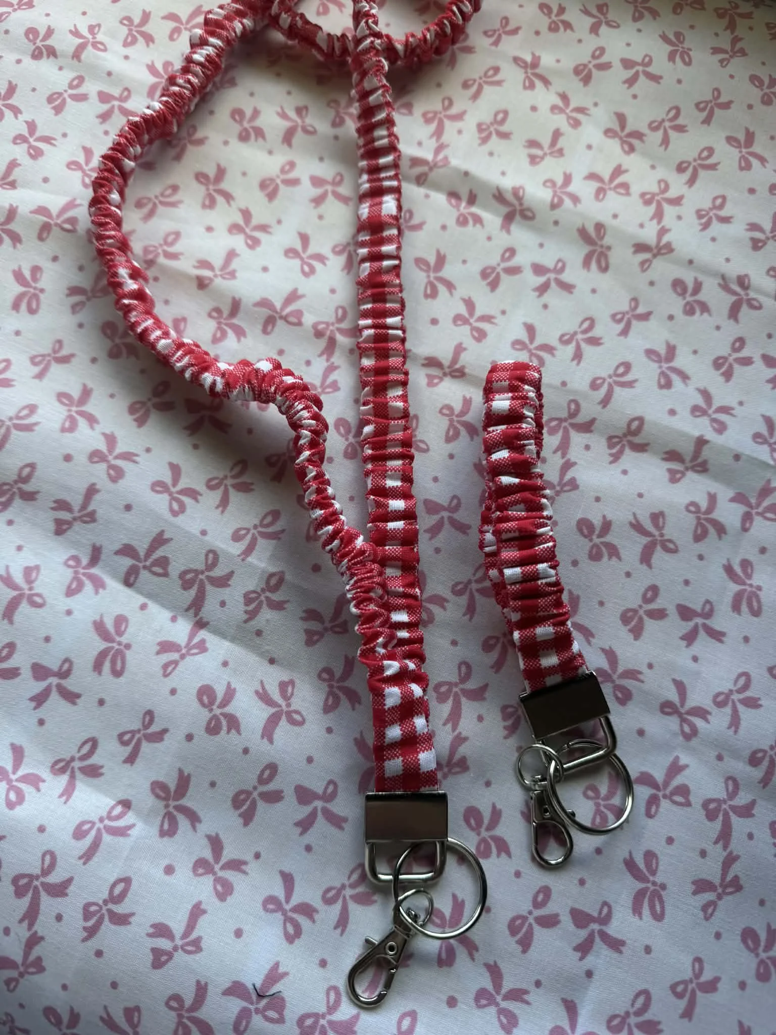 Red and white checkered elastic lanyards with metal clips on a pink and white bow patterned fabric background.