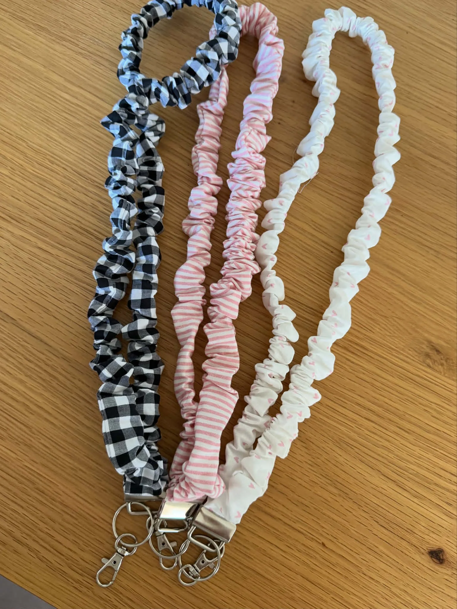 Three scrunchie keychains with metal clips on a wooden surface, in black and white checkered, pink and white striped, and white with pink hearts patterns.