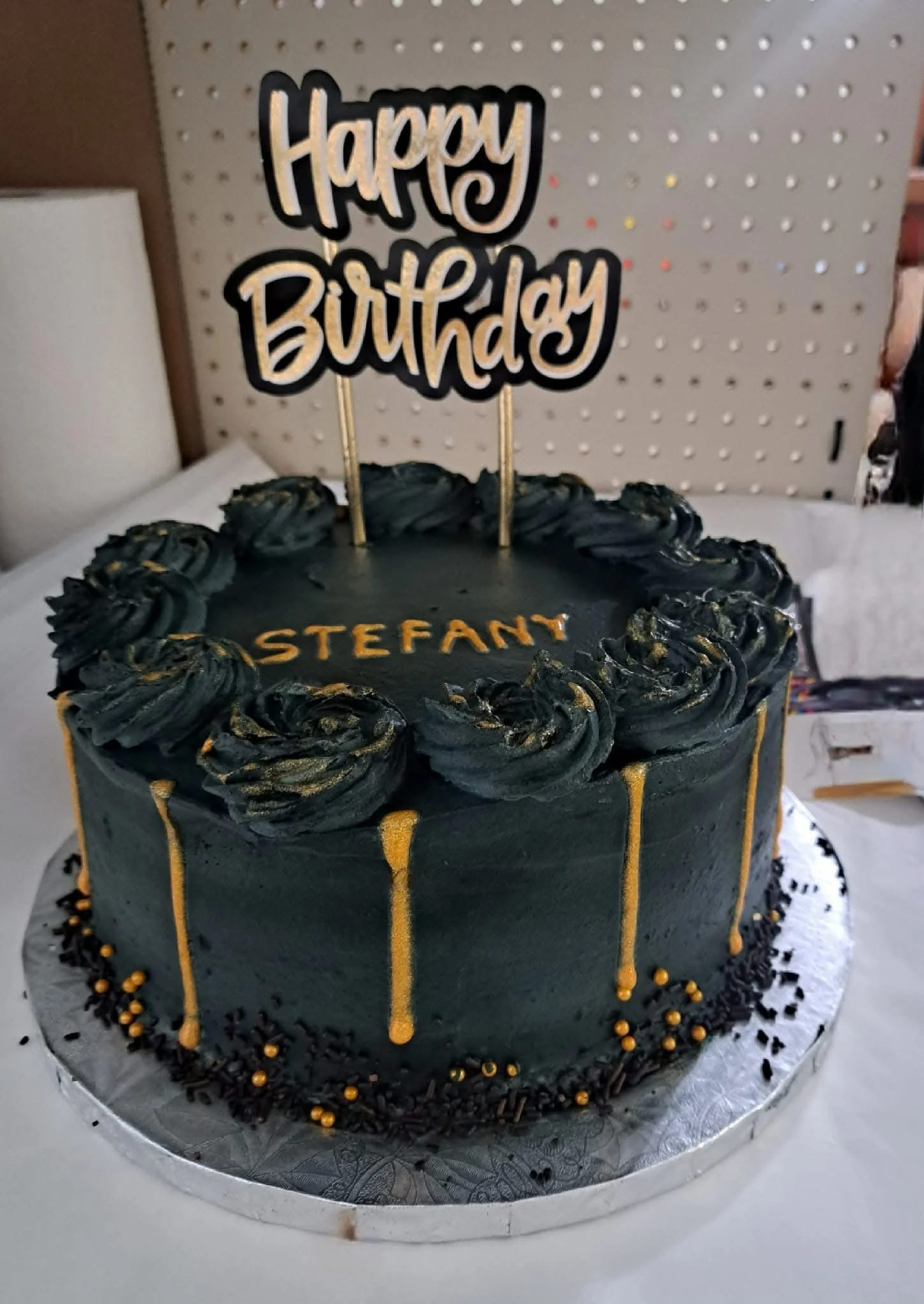 Black birthday cake with black and gold decorations, topped with a "Happy Birthday" sign, and the name 'Stefany' written on the cake.