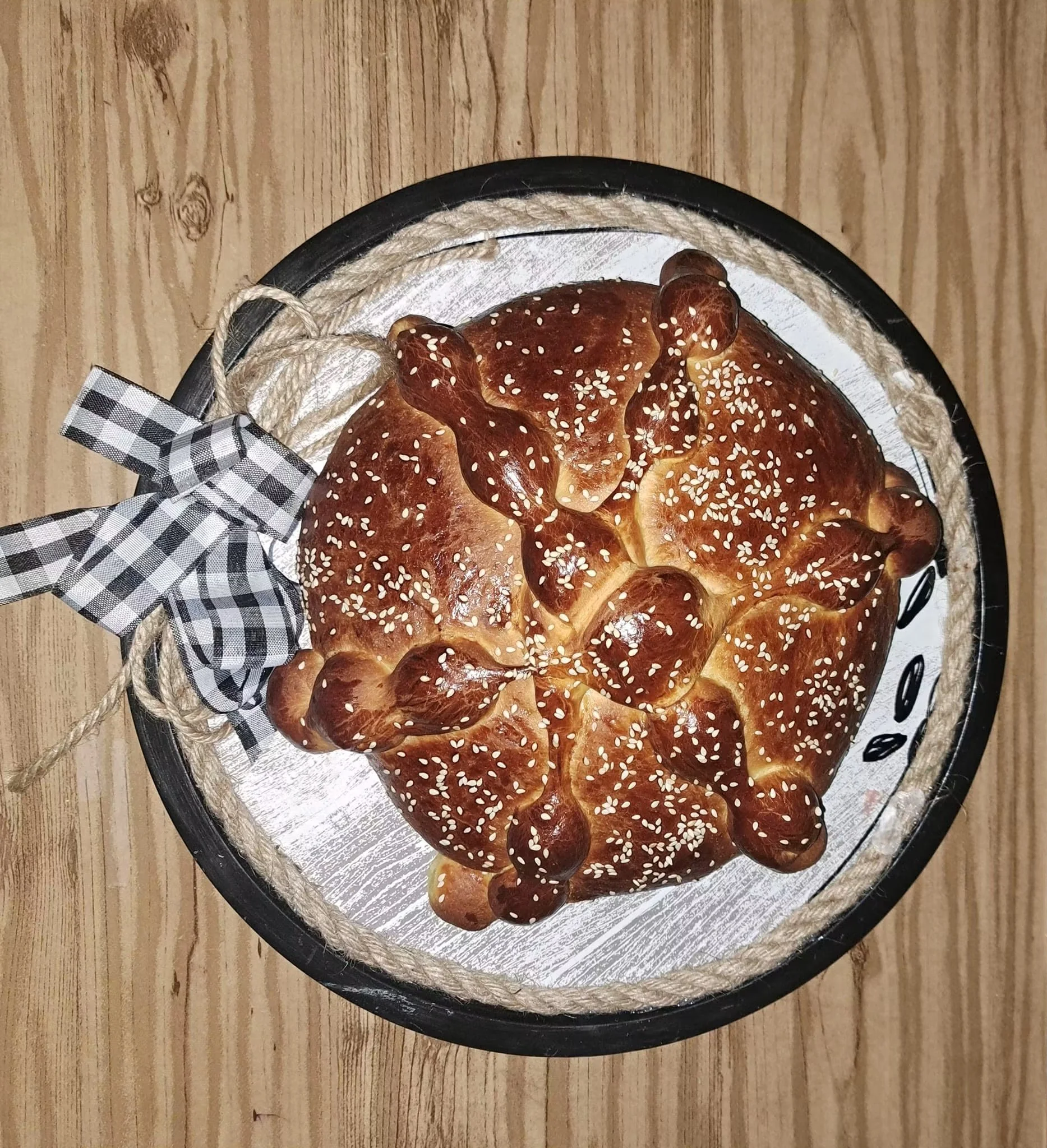 A round loaf of bread with sesame seeds on top, placed on a silver tray with a black border, decorated with a black-and-white checkered ribbon, on a wooden surface.