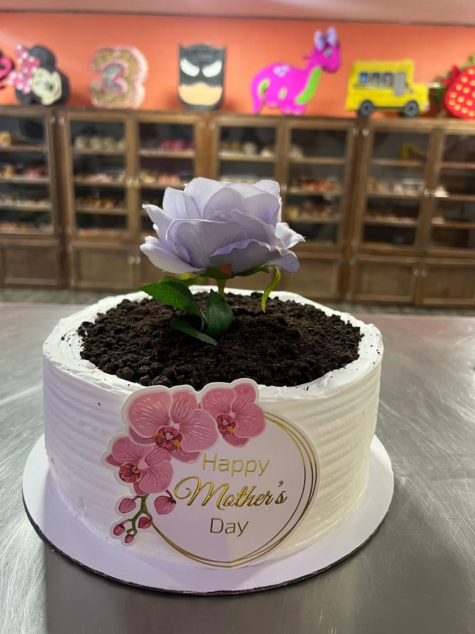 A round cake with white icing decorated with pink orchids and a label that reads "Happy Mother's Day". The cake is topped with a purple flower. The background shows a display case with various items and colorful wall decorations, including a pink uni