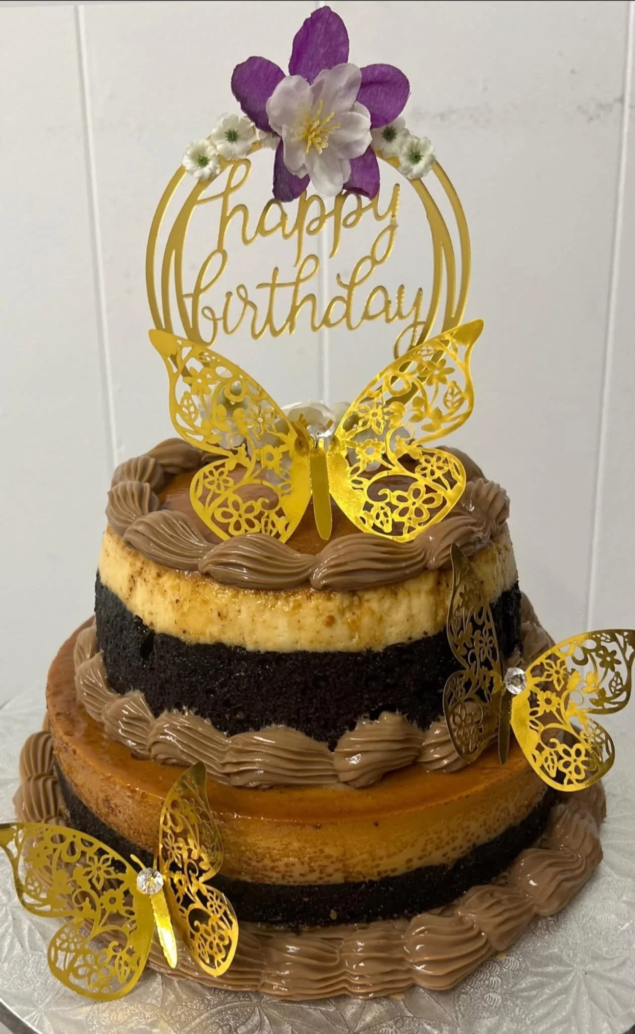 Multilayered birthday cake decorated with gold butterfly toppers, chocolate frosting, and a topper that reads 'happy birthday' with purple and white flowers.