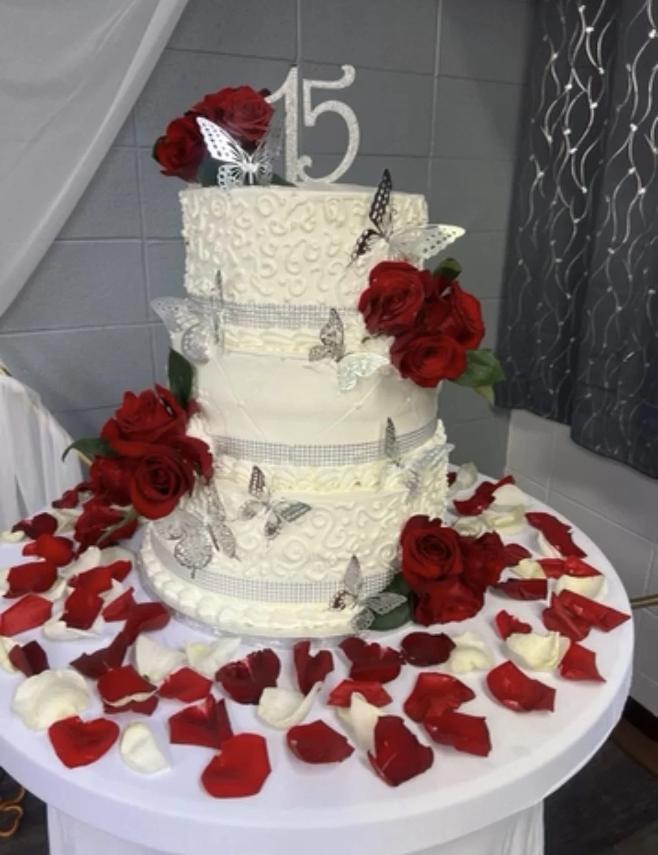 A three-tier white wedding cake decorated with red roses, silver butterflies, and a '15' topper, surrounded by red and white rose petals on a round white table.