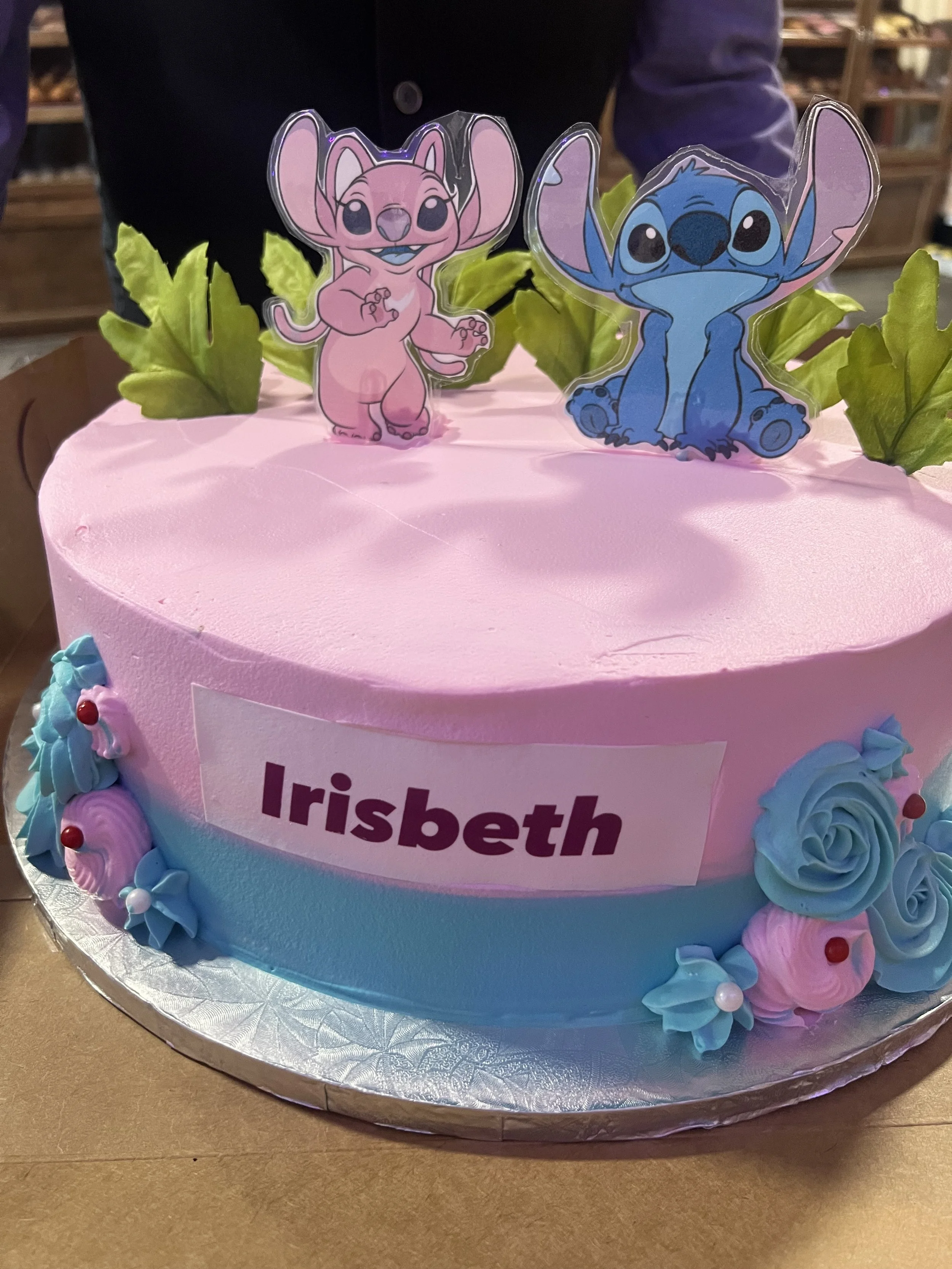 A birthday cake decorated in pink and blue with colorful flowers and cartoon character toppers of Stitch and Angel from Lilo & Stitch.