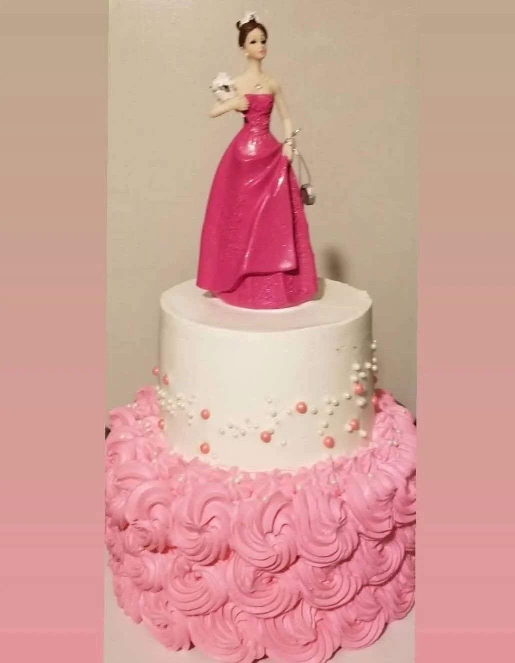 A birthday cake with two tiers. The top tier has a figurine of a woman in a pink ball gown, holding a white flower, standing on a white base. The bottom tier is decorated with pink rosette-style icing and small pink and white spherical decorations.