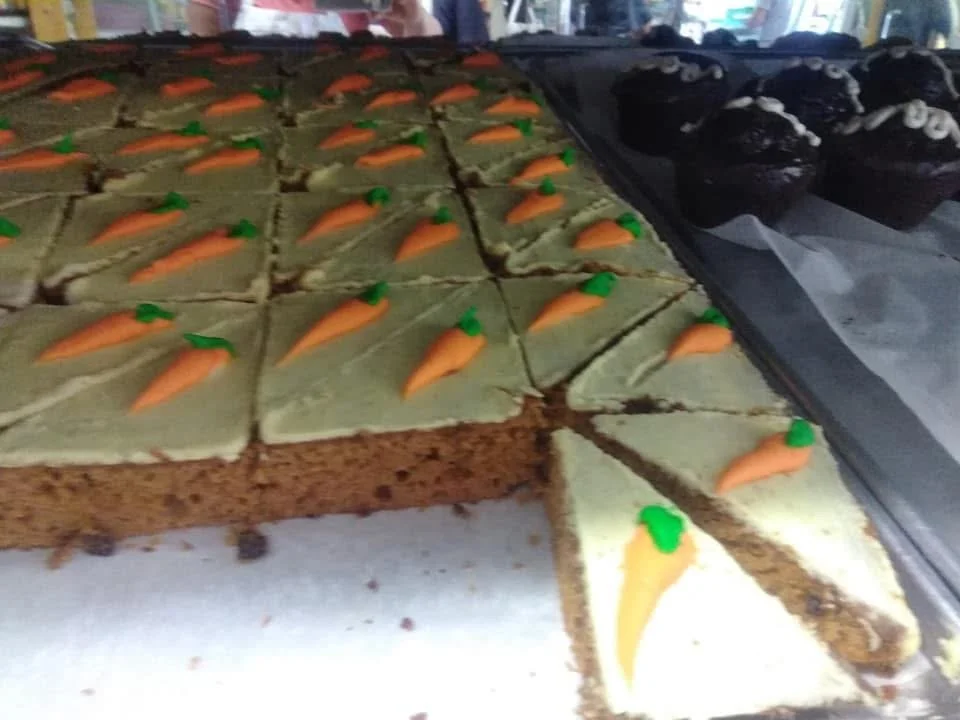 A tray of carrot cake squares decorated with small carrot-shaped icing toppers and a few black cupcakes with white decorations in the background.
