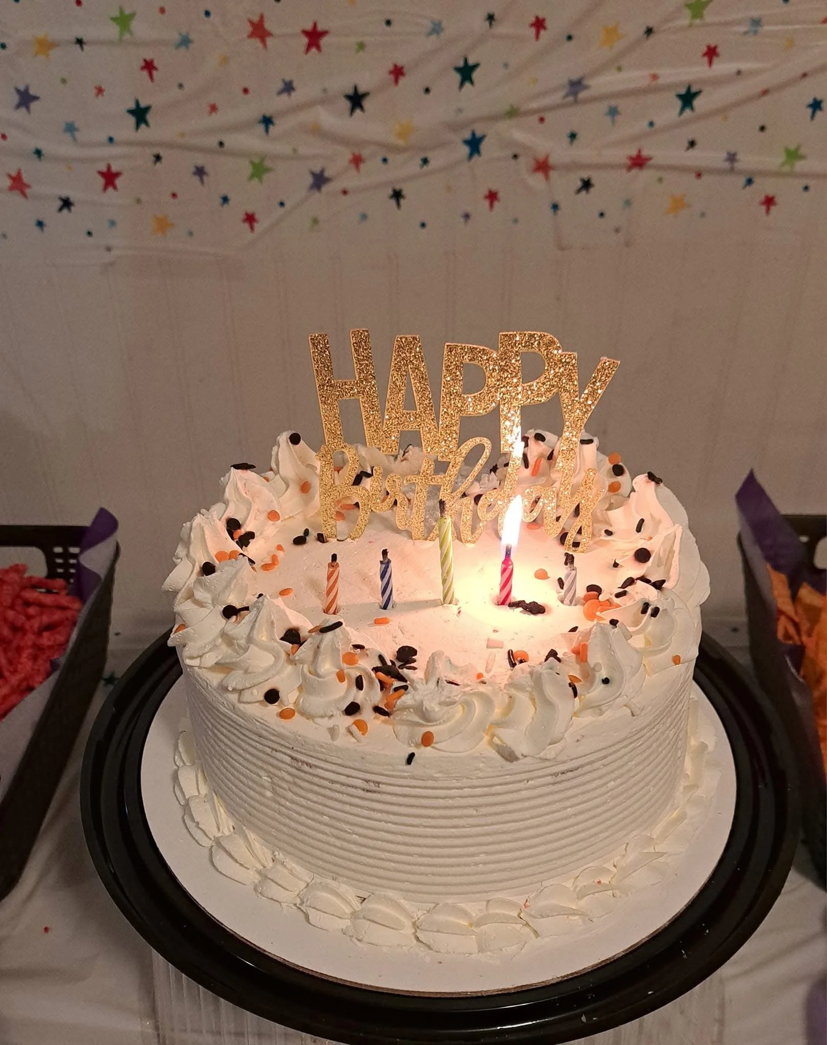 A birthday cake decorated with white frosting, black and orange sprinkles, and a cake topper that says 'Happy Birthday' in gold glitter. There are five lit candles on the cake, each with a different color stripe.