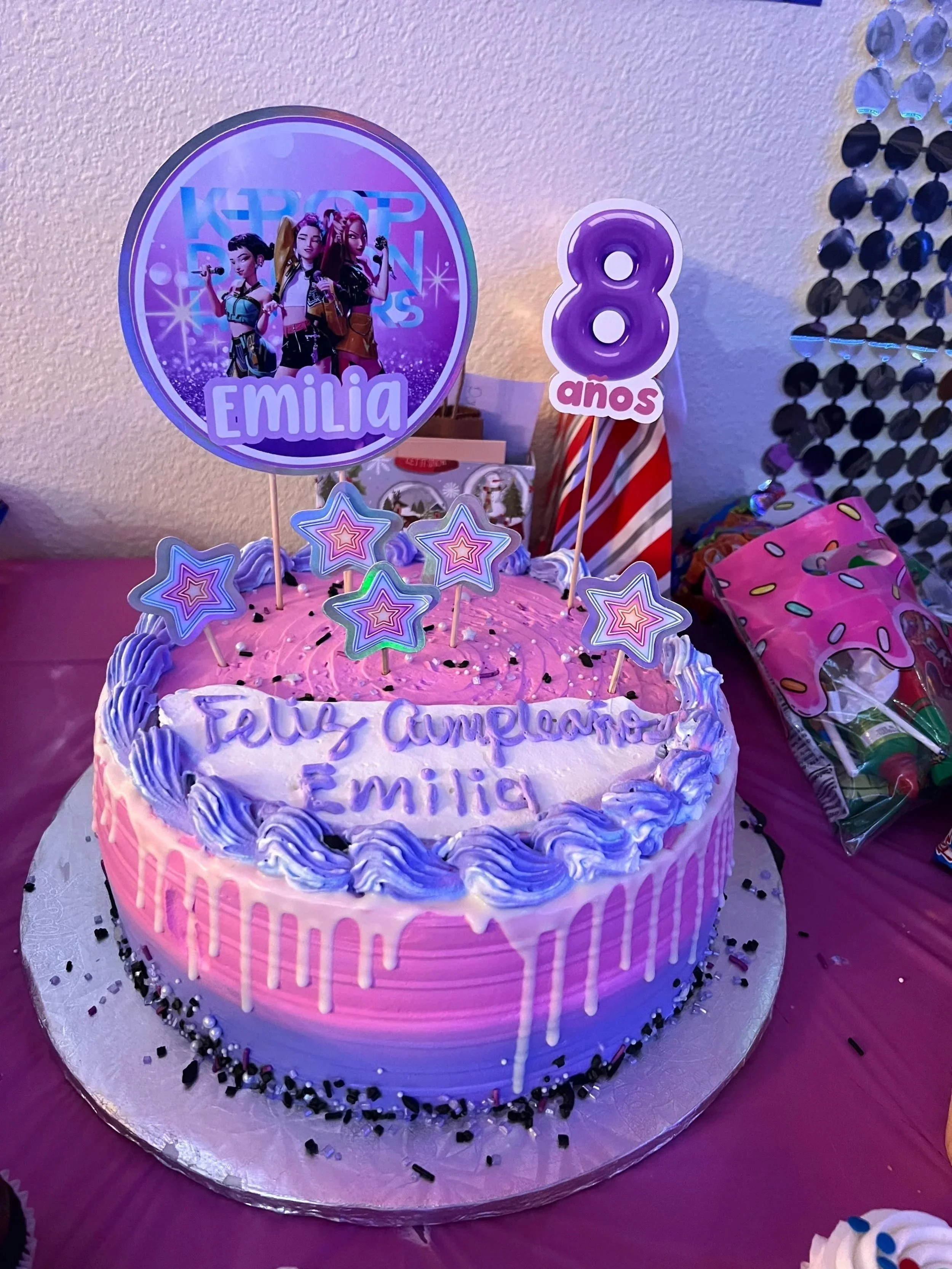 A birthday cake with pink and purple icing decorated with star-shaped and round-shaped toppings, with a large themed topper featuring three animated characters and the text 'Emilia', and a smaller sign displaying '8 años'.