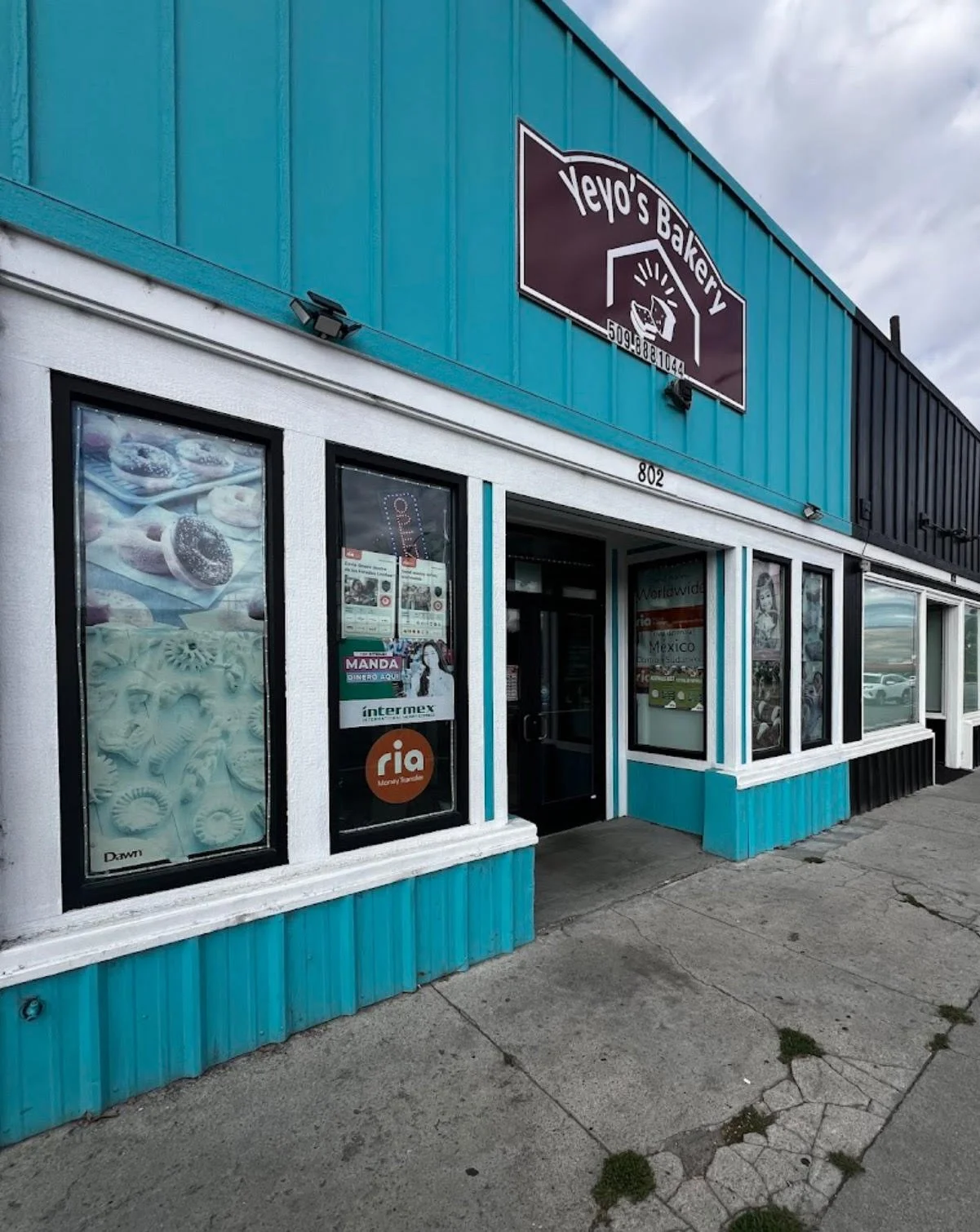 The exterior of a bakery shop with large display windows showcasing baked goods, a sign reading 'Yeyo's Bakery' above the entrance, and various stickers on the door. The building is painted with turquoise and white accents.