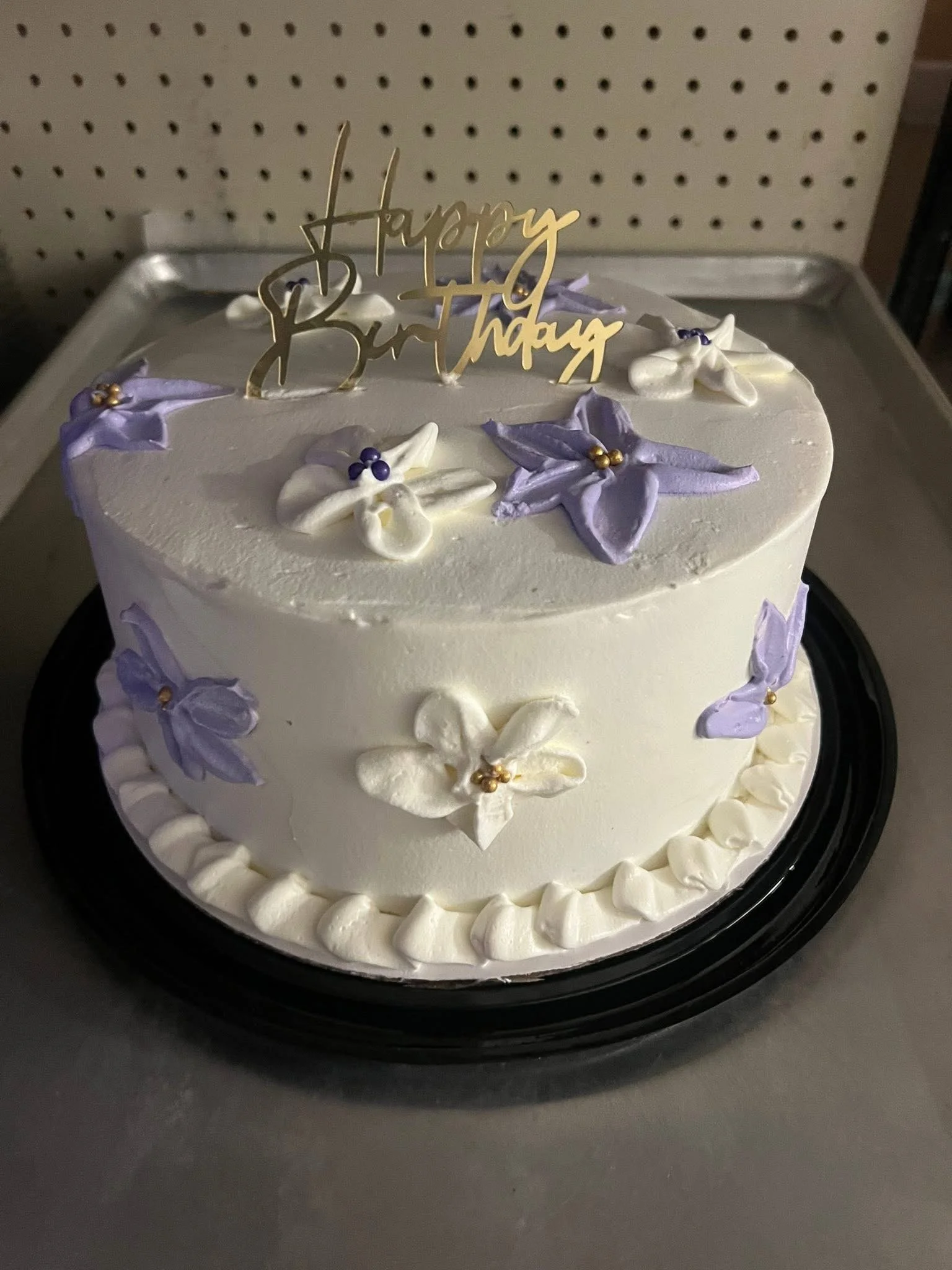 A white frosted birthday cake decorated with purple and white star-shaped flowers, gold bead accents, and a gold 'Happy Birthday' topper.