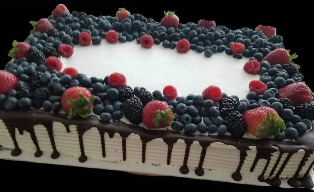 A rectangular cake decorated with an assortment of fresh berries, including strawberries, blueberries, blackberries, raspberries, and blackberries, on top and along the sides, with a white cream filling and chocolate drizzle on the edges.