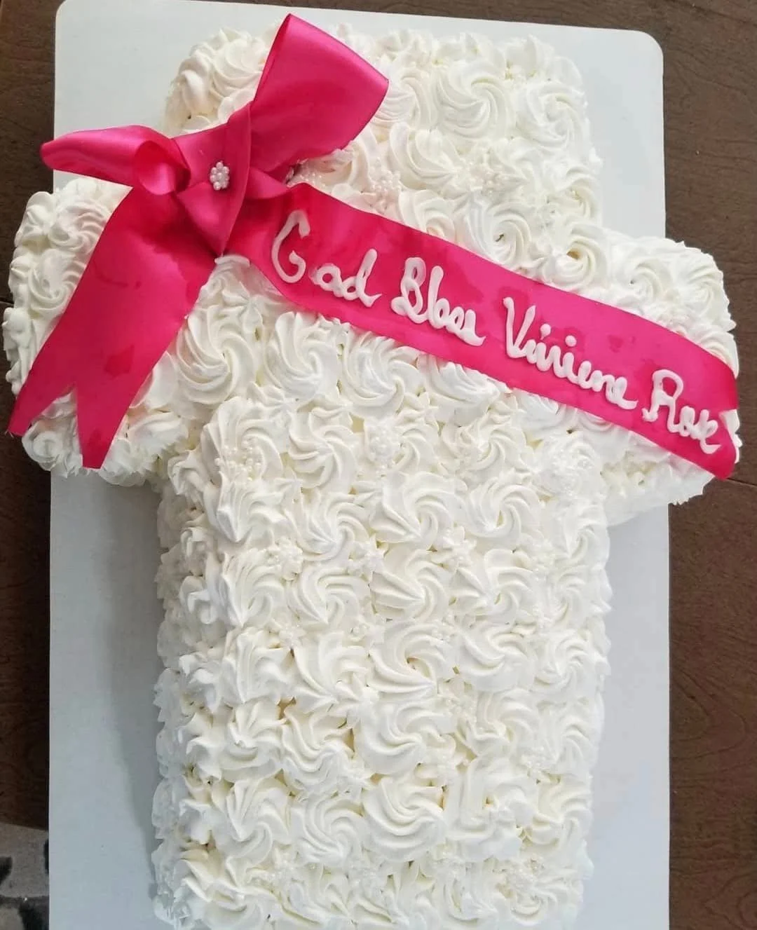 White frosted sheet cake in the shape of the number 1 with a pink ribbon and bow on top. The ribbon has the message 'God Bless Viminee Raw' written on it in white icing.