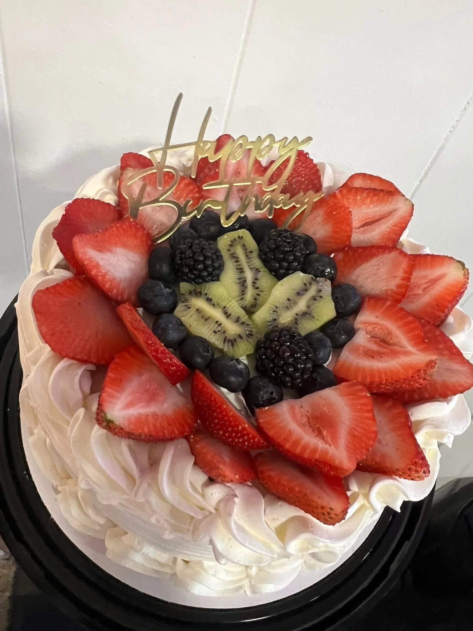 A cake decorated with sliced strawberries, blueberries, blackberries, and kiwi slices, topped with a golden 'Happy Birthday' sign.